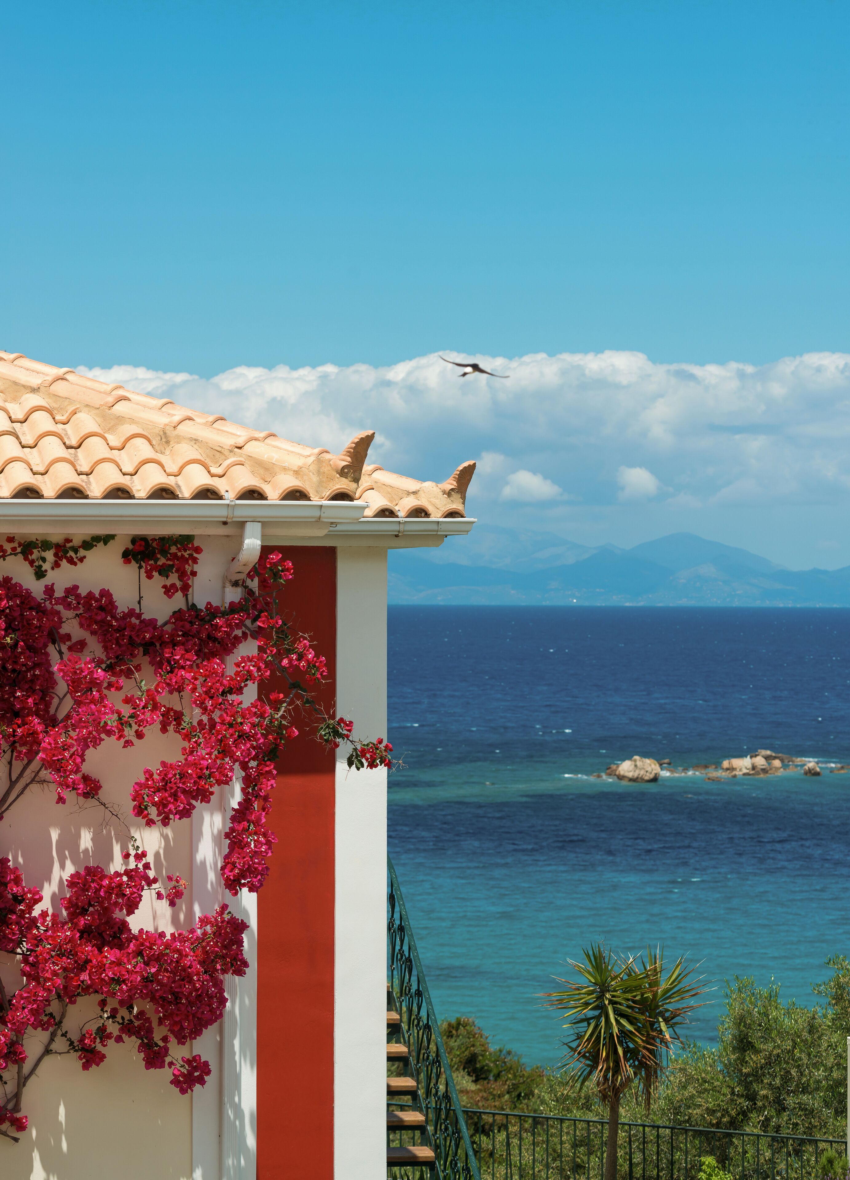 Ein Küstenhaus mit Ziegeldach, einem Balkon mit roten Blumen und Blick auf das Meer und die Berge.