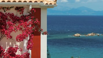 Ein Küstenhaus mit Ziegeldach, einem Balkon mit roten Blumen und Blick auf das Meer und die Berge.