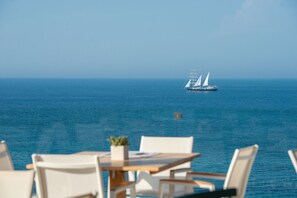 Uma mesa e cadeiras viradas para a vista do mar, com um veleiro ao longe.
