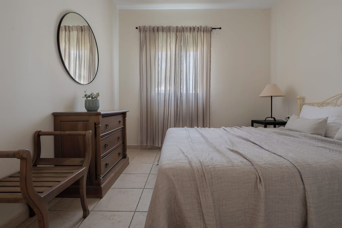 A bedroom with a bed, a wooden bench, a dresser, a mirror, a lamp, and a window with curtains.