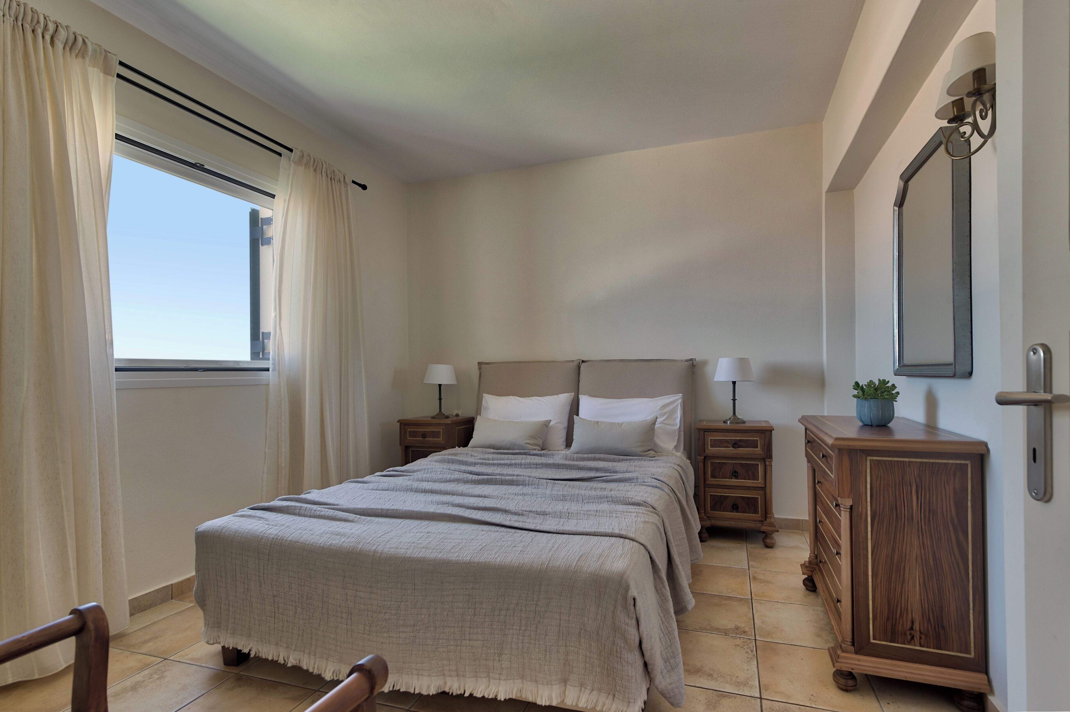 Une chambre à coucher avec un lit, des tables de chevet, une commode, un miroir et une fenêtre avec des rideaux.
