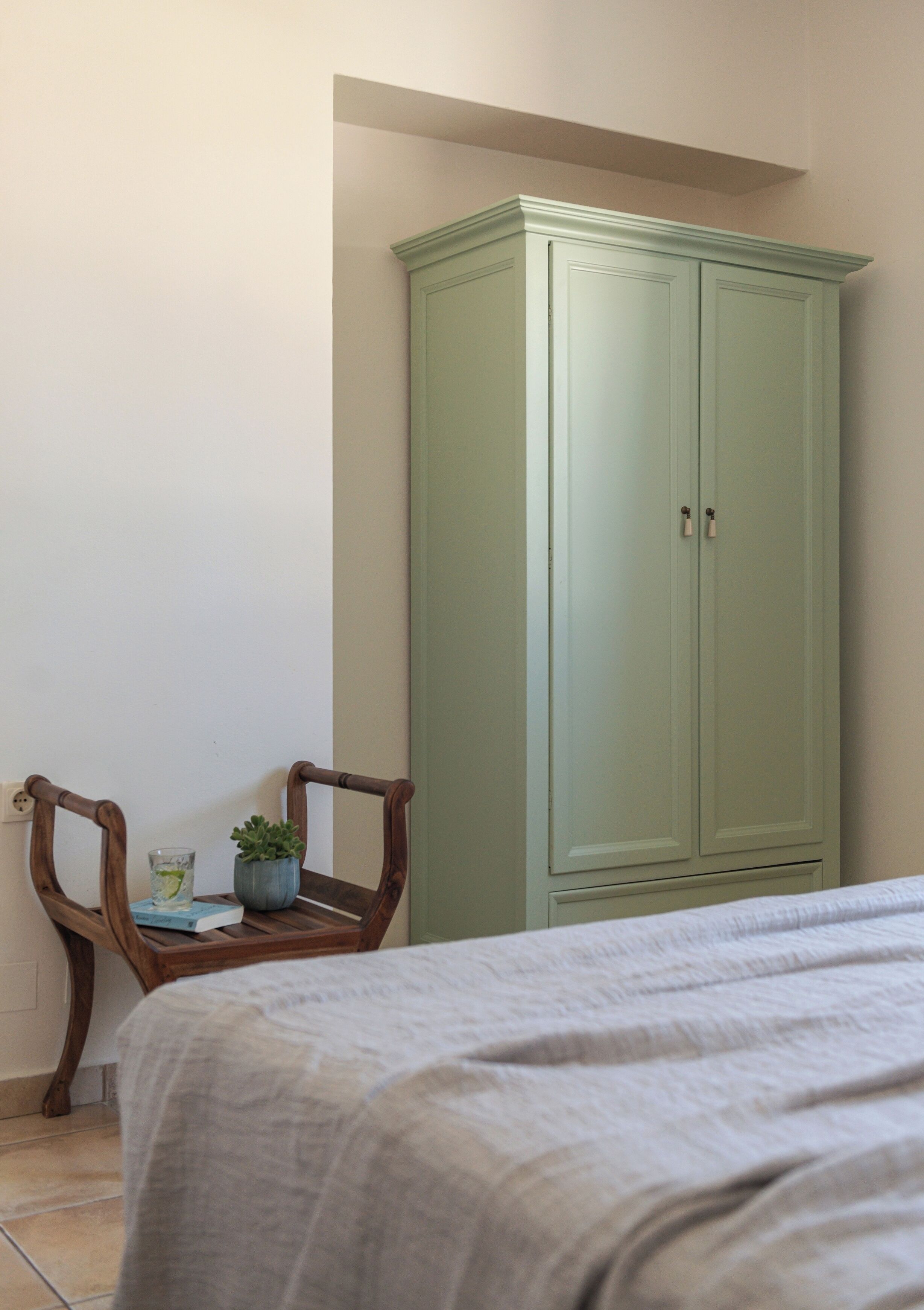 Un dormitorio con una cama, una silla de madera, una mesita con una planta y un armario verde.