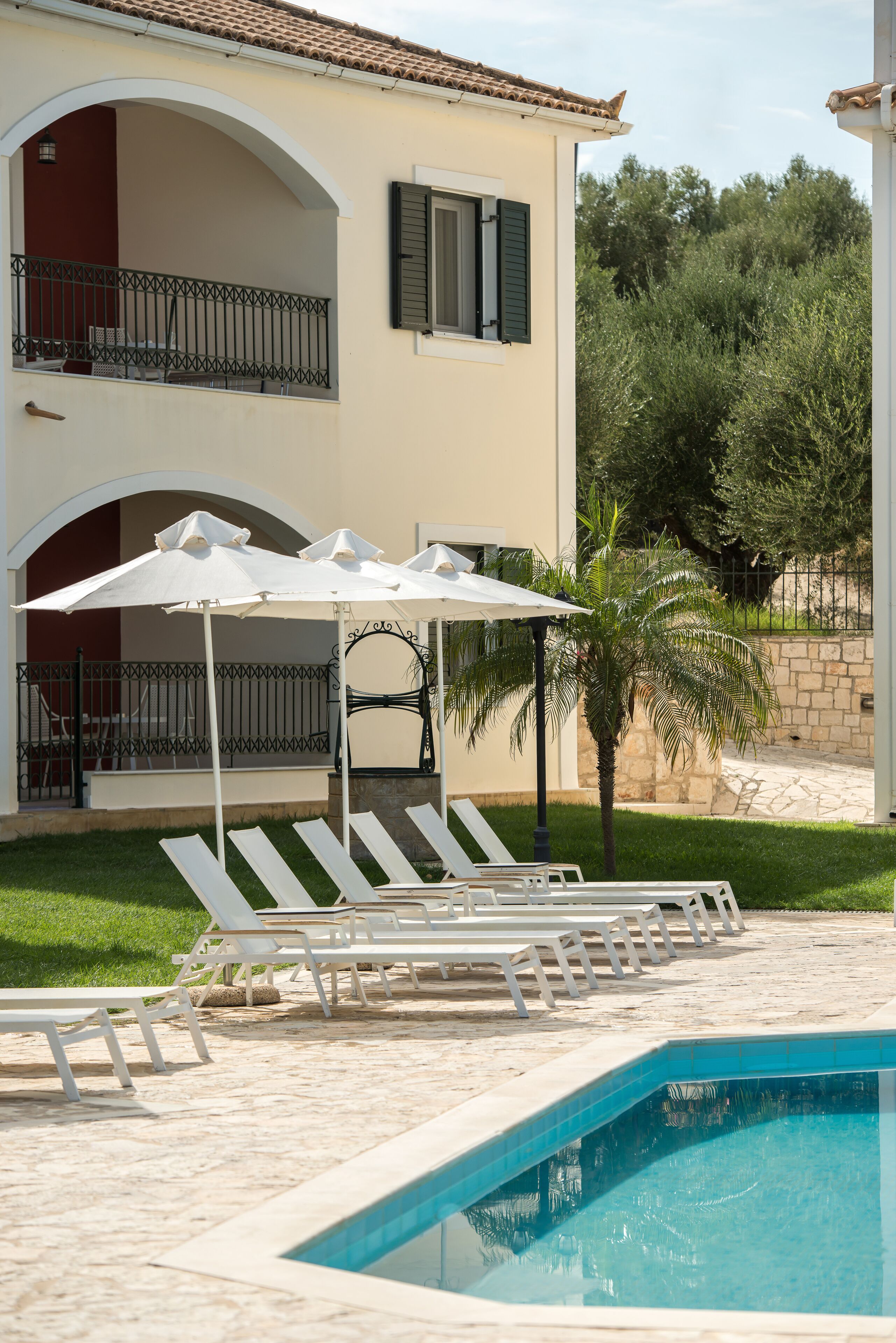 Un espace aménagé au bord de la piscine, avec des chaises longues et des parasols, une piscine, et un bâtiment doté d’un balcon.