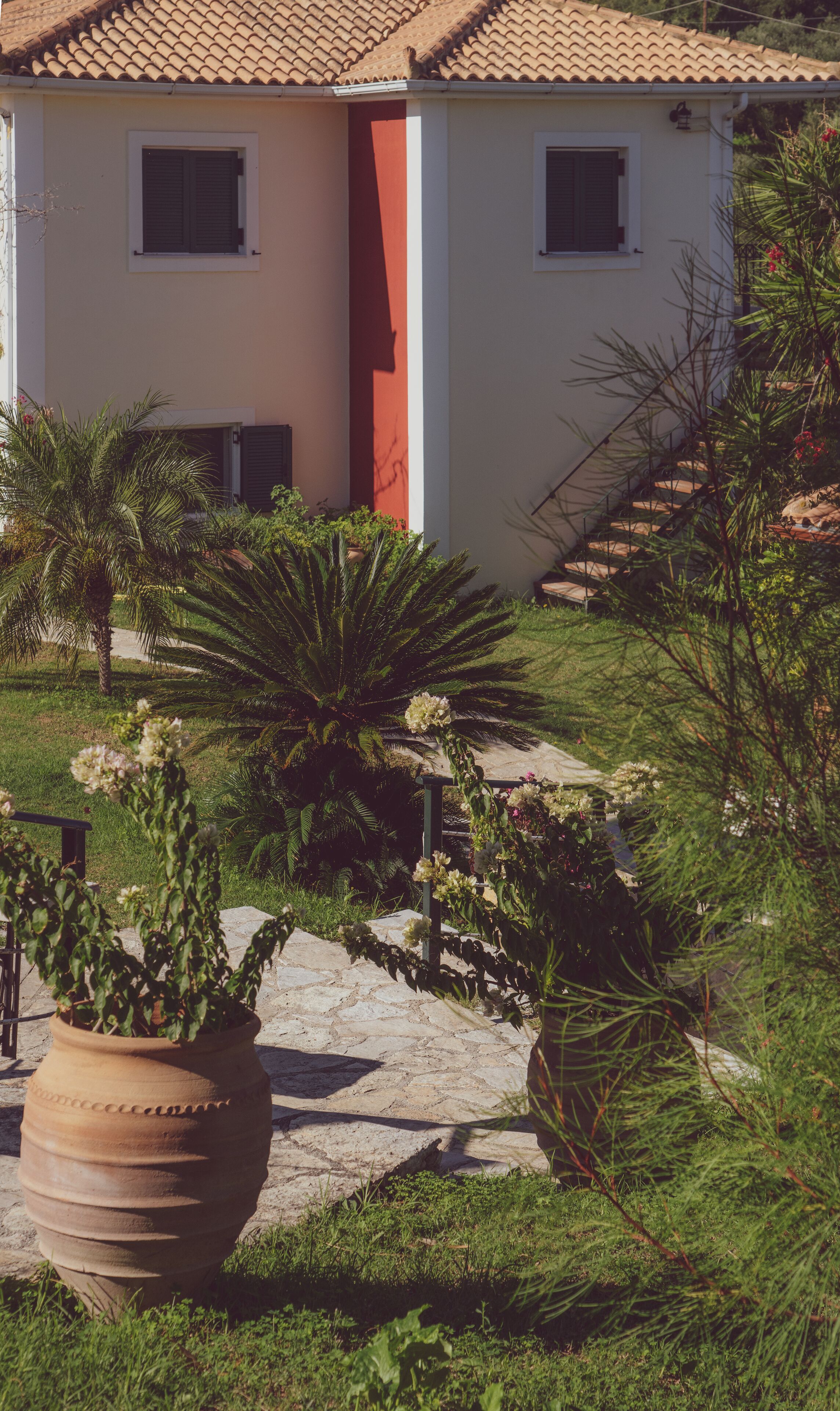 A house with a tiled roof, a red door, and a garden with various plants and a large pot.