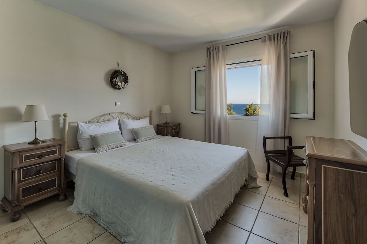 A neatly arranged bedroom with a bed, bedside tables, a chair, and a view of the sea through the window.
