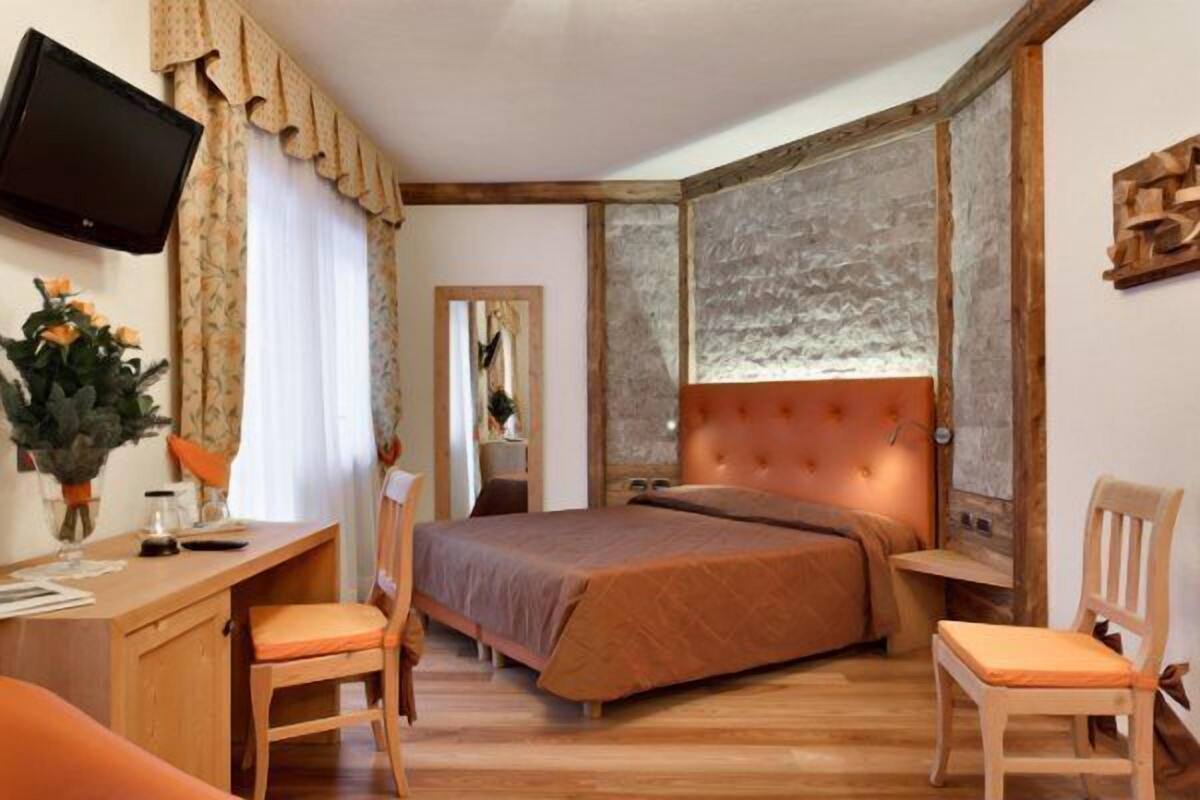 A bedroom with a wooden headboard, a bed with a brown bedspread, a desk with a chair, and a television mounted on the wall.