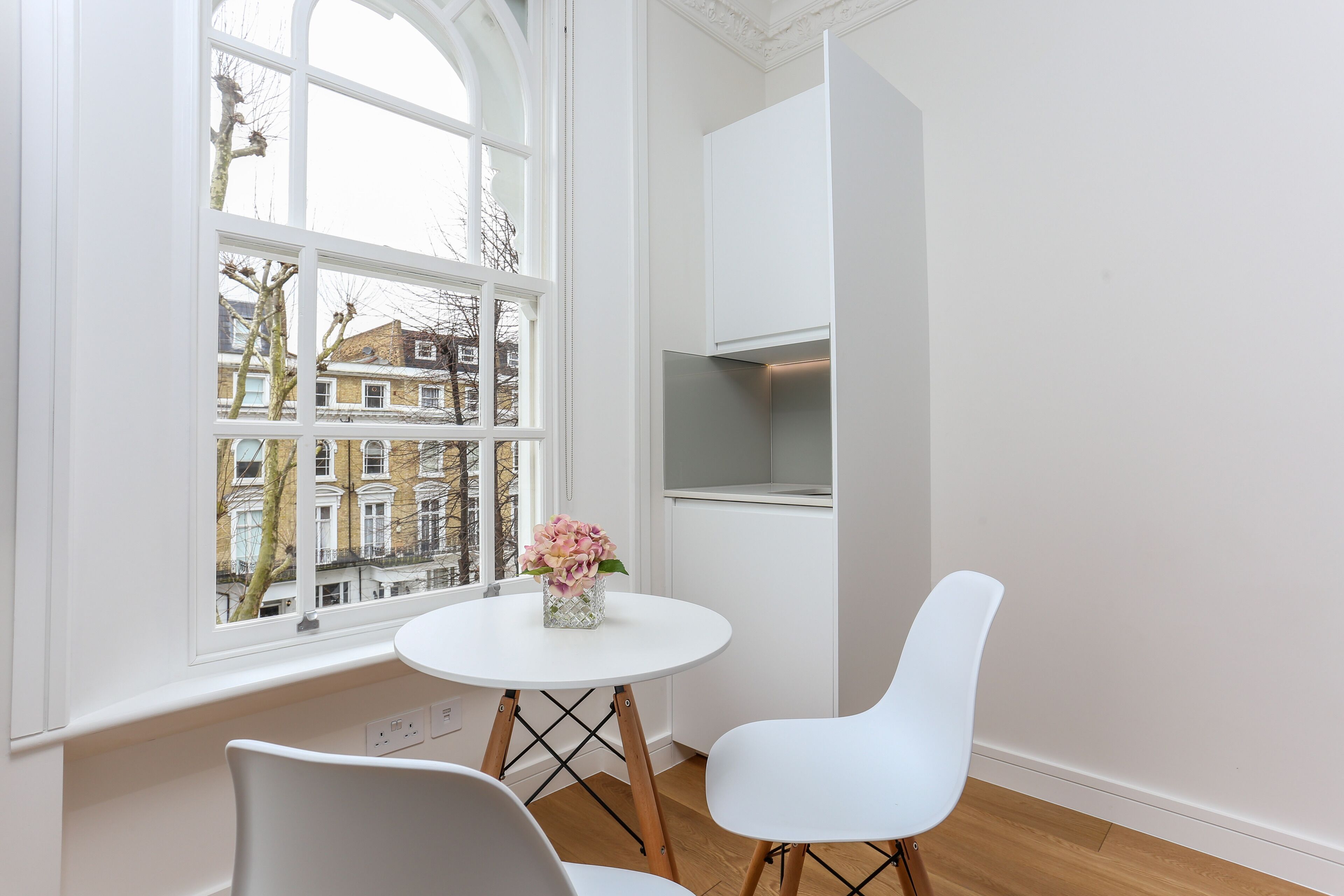 A modern room with a round white table, two white chairs, a vase with flowers, and a view of buildings through a large window.
