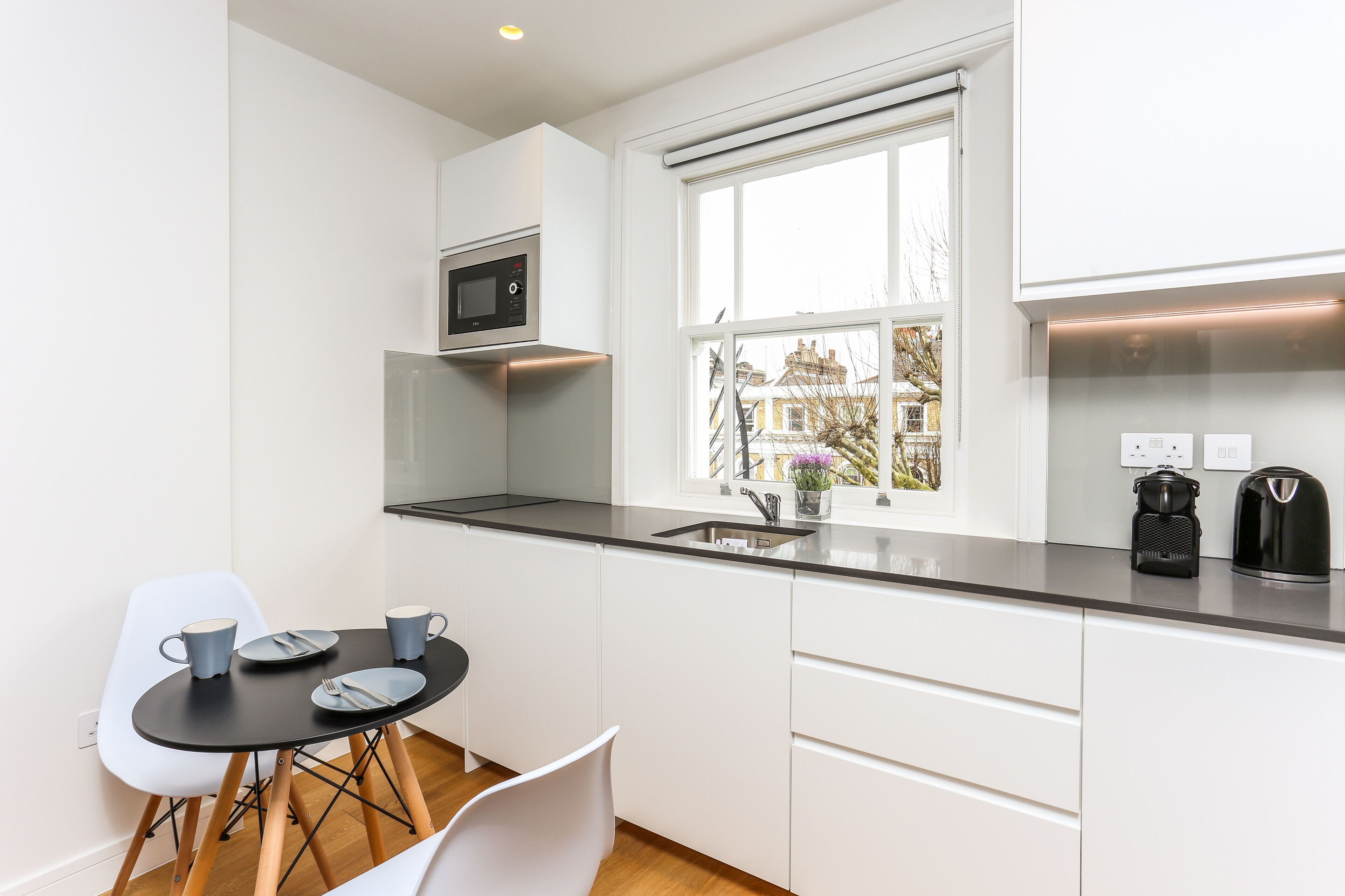 A compact kitchen with white cabinets, a black countertop, a microwave, a coffee maker, and a window with a view of buildings.