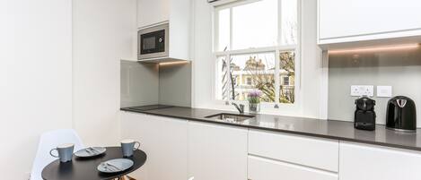 A compact kitchen with white cabinets, a black countertop, a microwave, a coffee maker, and a window with a view of buildings.
