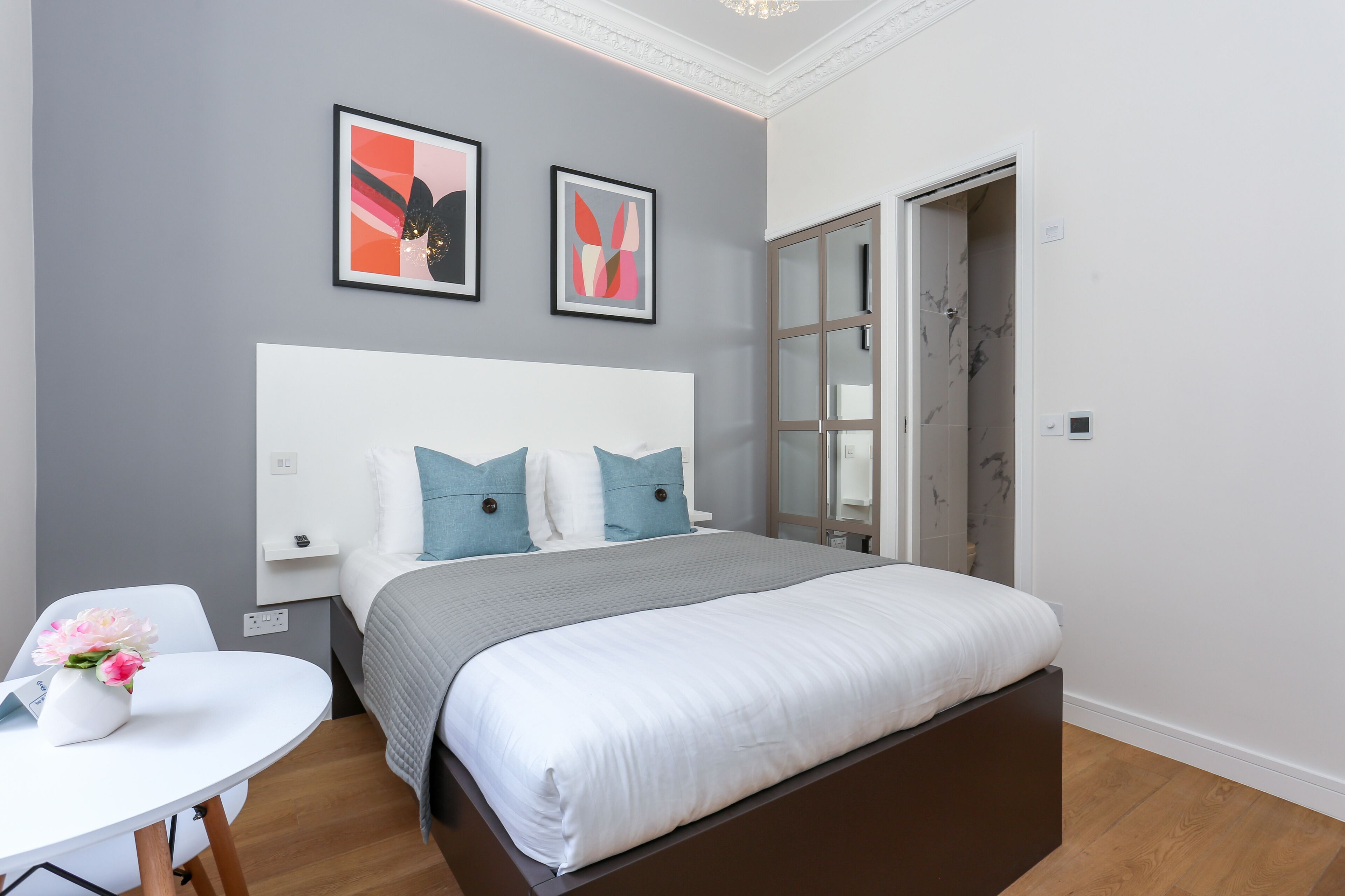 A modern bedroom with a large bed, a bedside table, and a chair. There are two framed artworks on the wall above the bed.