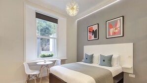 A modern bedroom with a large bed, bedside tables, a small desk, and a window with a view of greenery. - Inverness Terrace Serviced Apartments by Concept Apartments (London)