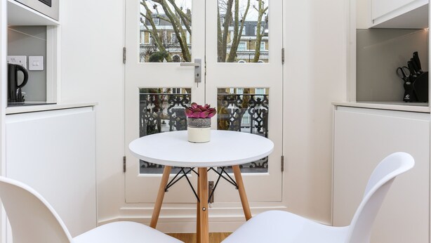 A room with a white round table, two white chairs, a potted plant, and a view through a glass door. - Inverness Terrace Serviced Apartments by Concept Apartments (London)