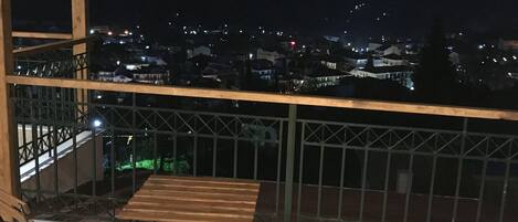 A balcony with two wooden tables and chairs, a metal railing, and a view of a cityscape at night.