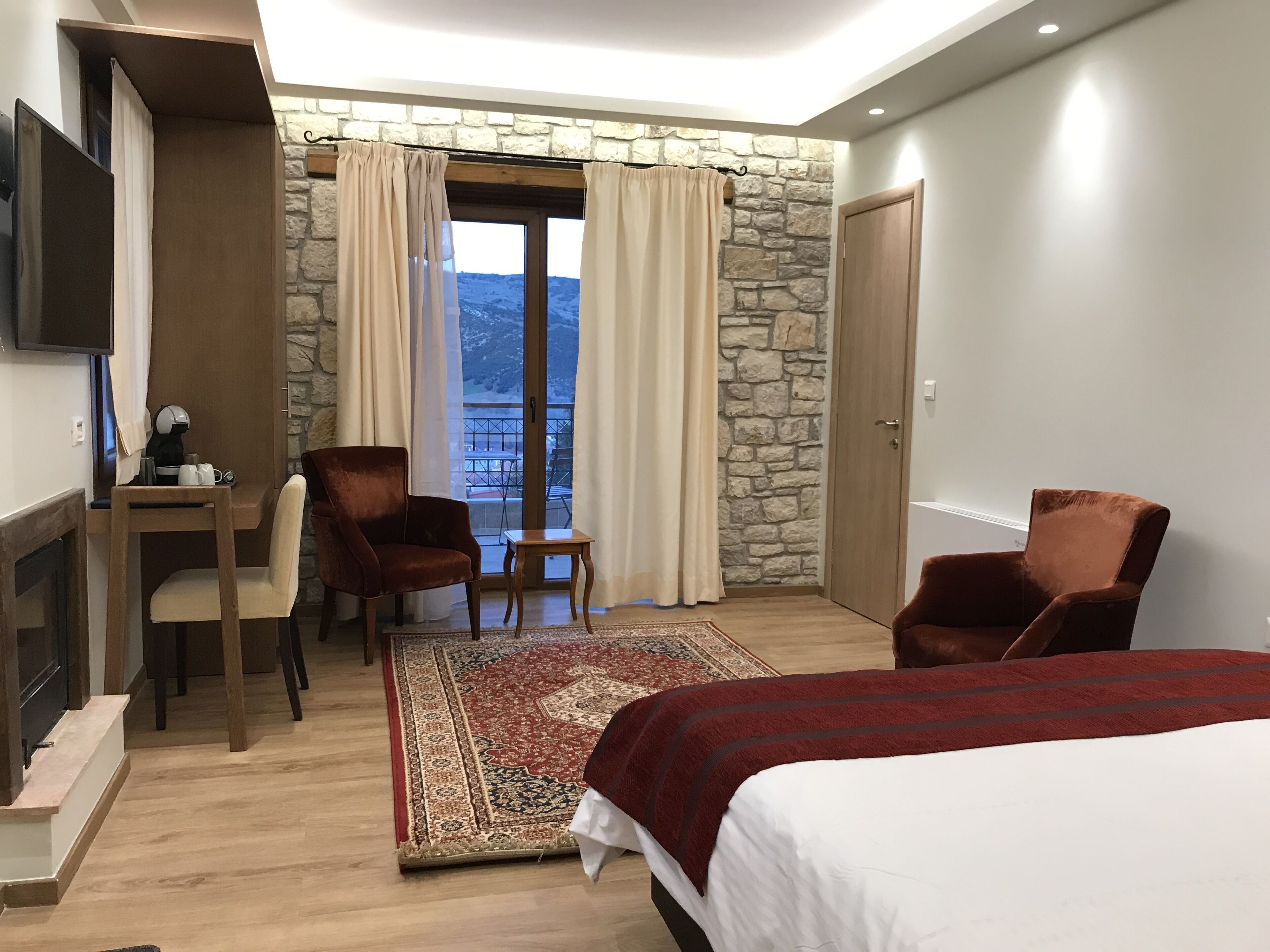 A hotel room with a stone wall, a balcony with a view, a desk with a chair, a bed, and a TV mounted on the wall.