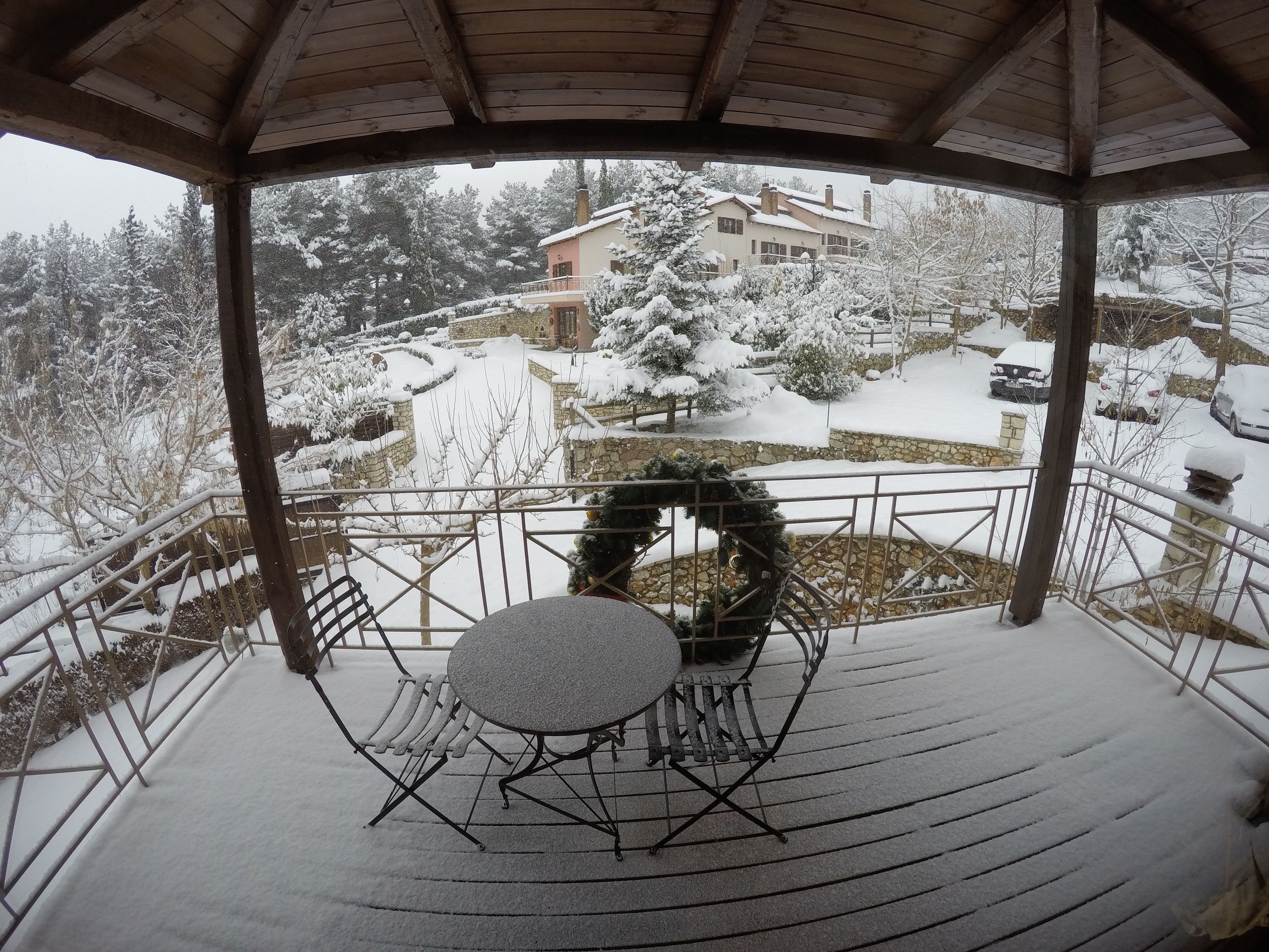 A covered outdoor area with a table and chairs, a stone wall, and a view of a snow-covered building and trees.