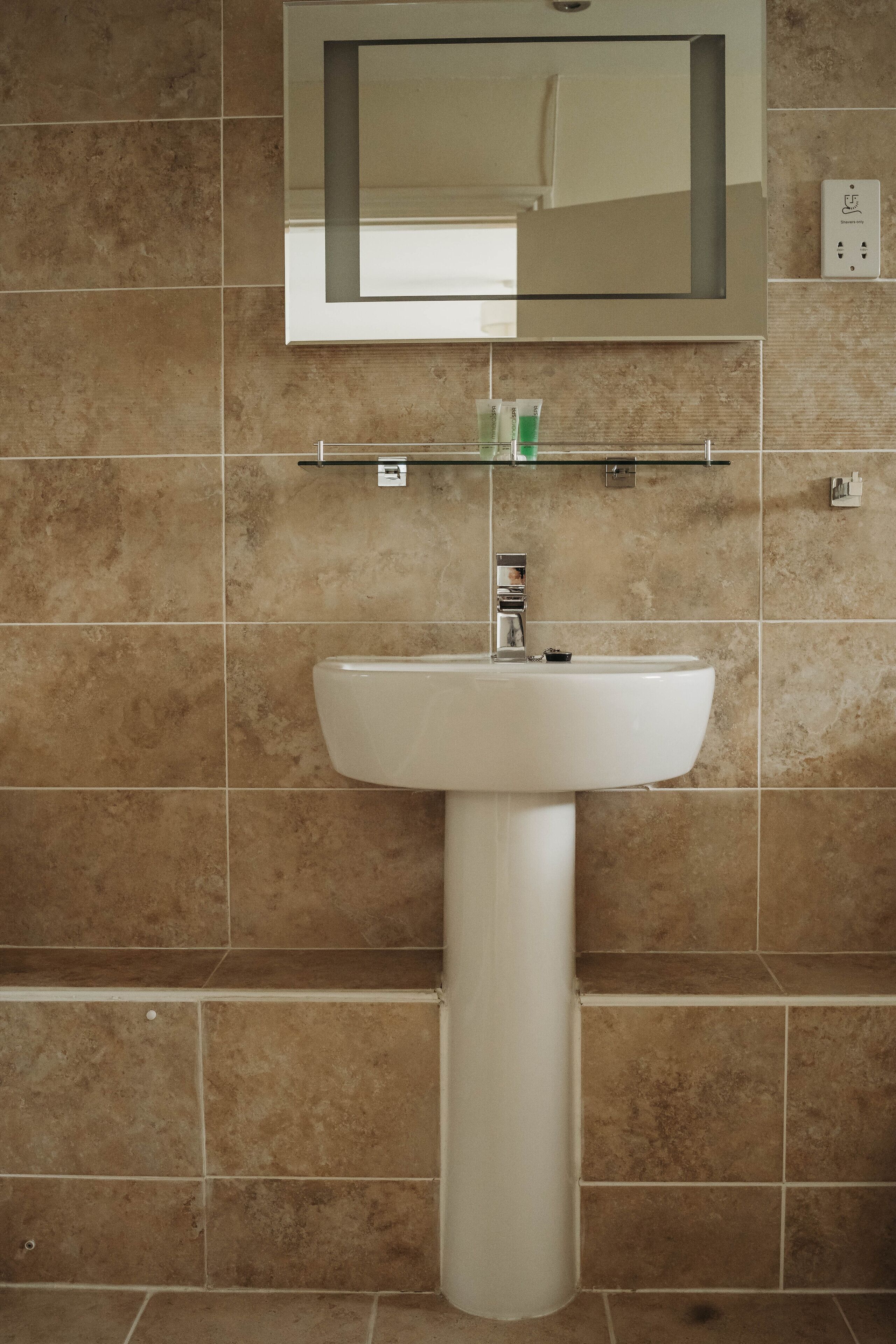 Un baño con un lavamanos blanco de pedestal, un espejo encima y un estante con artículos de aseo.