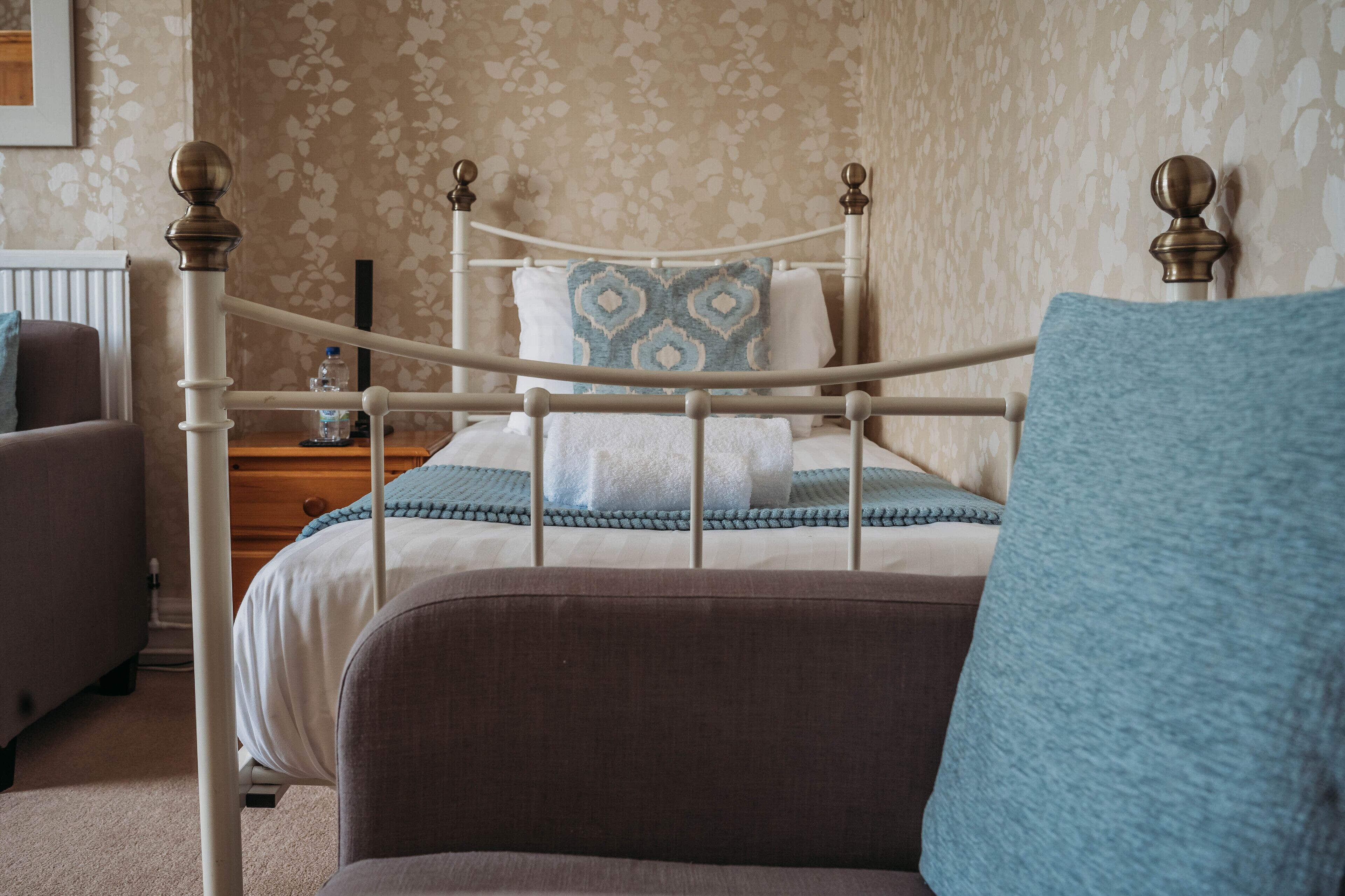 A neatly made bed with a patterned pillow, a bedside table with a bottle, and a chair with a cushion.