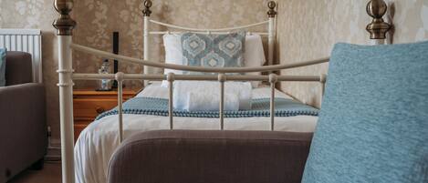 A neatly made bed with a patterned pillow, a bedside table with a bottle, and a chair with a cushion.