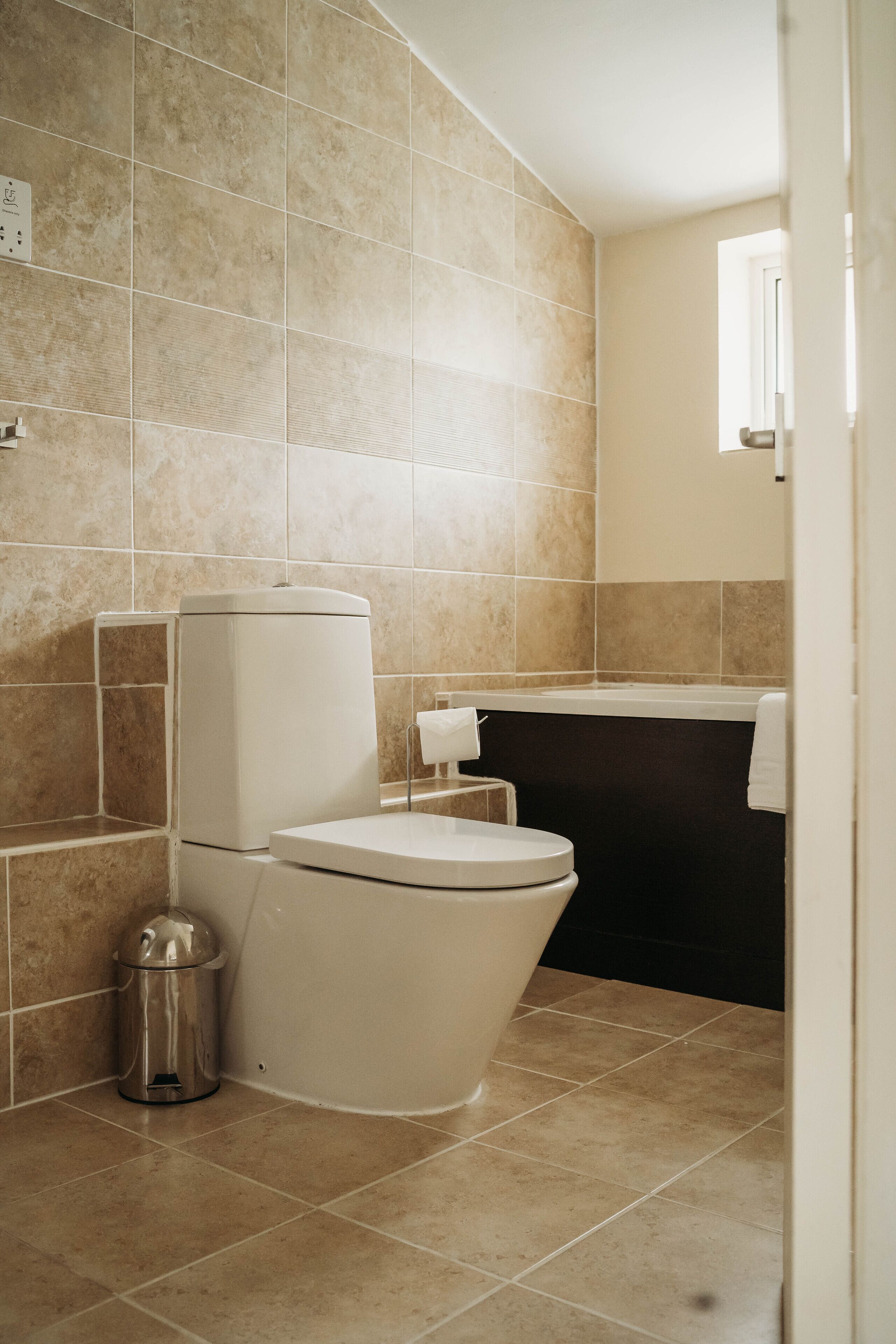 Un baño con azulejos color beige, un inodoro, un cubo de basura y una bañera.