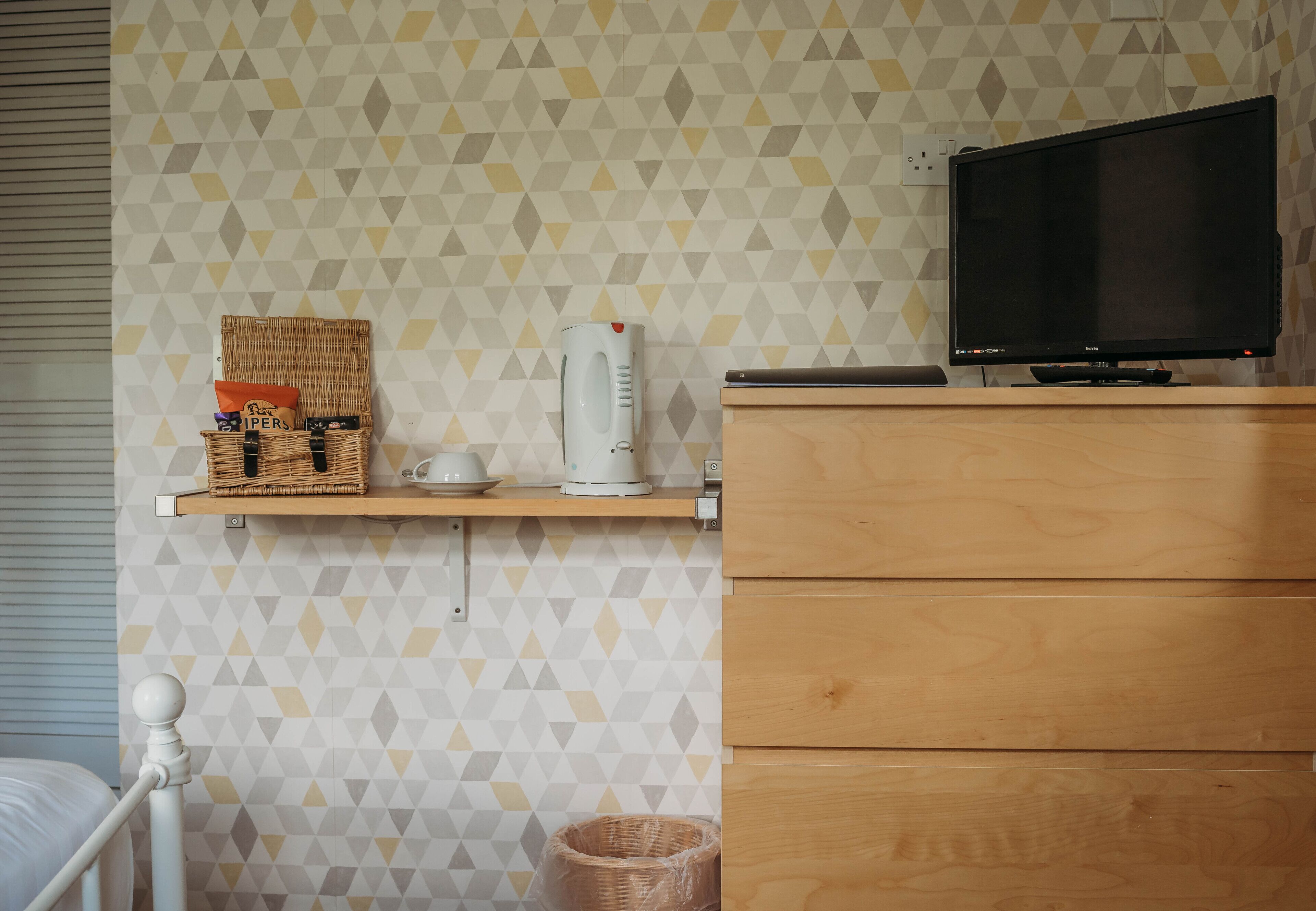 A bedroom with a bed, a wooden dresser, a television, a shelf with a basket and a kettle.