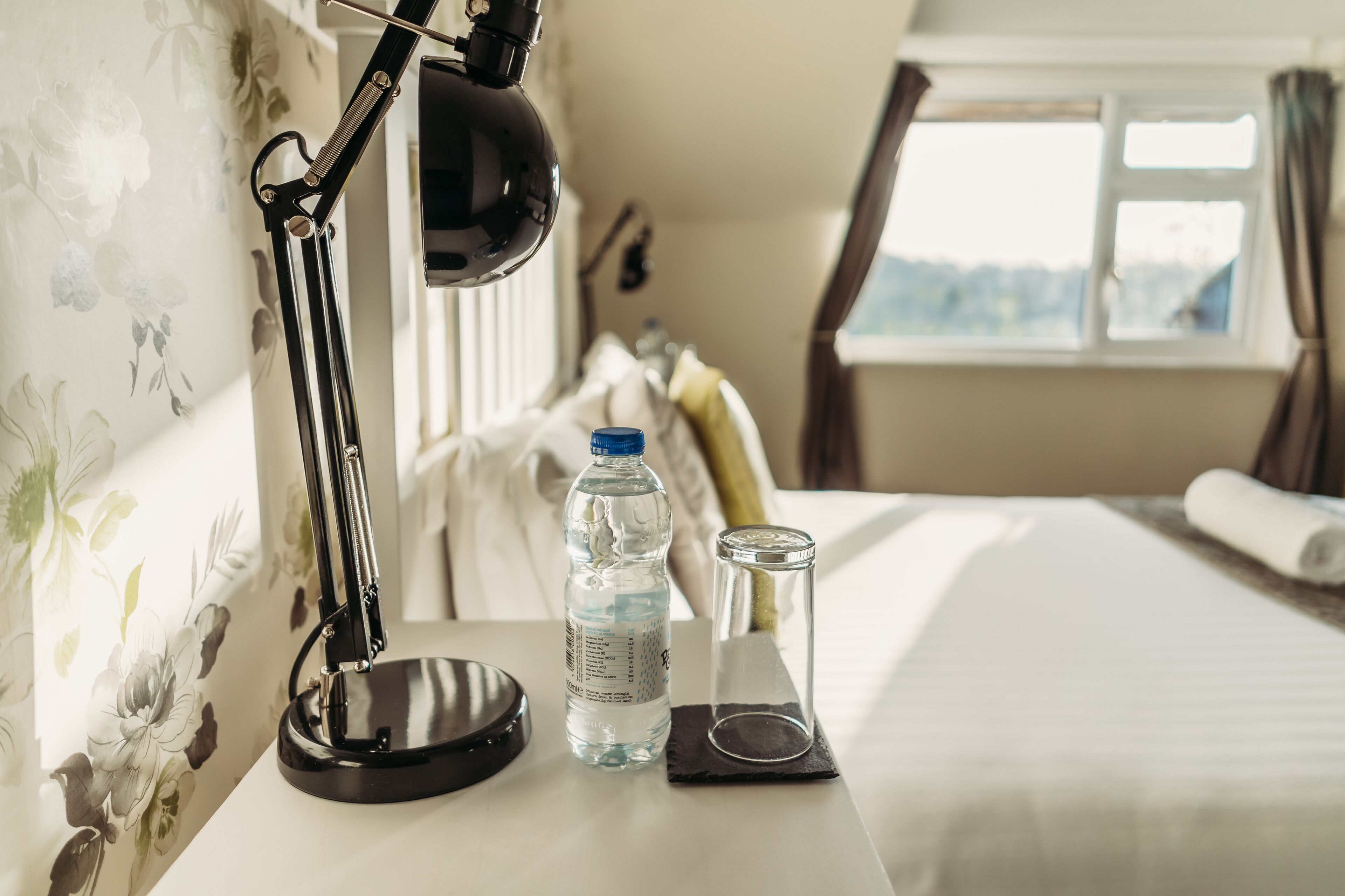 A neatly arranged hotel room with a desk, a water bottle, a glass, and a lamp.