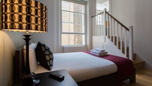 A hotel room with a bed, a nightstand with a lamp, a window, and a staircase. - Princes Square Serviced Apartments by Concept Apartments (London)