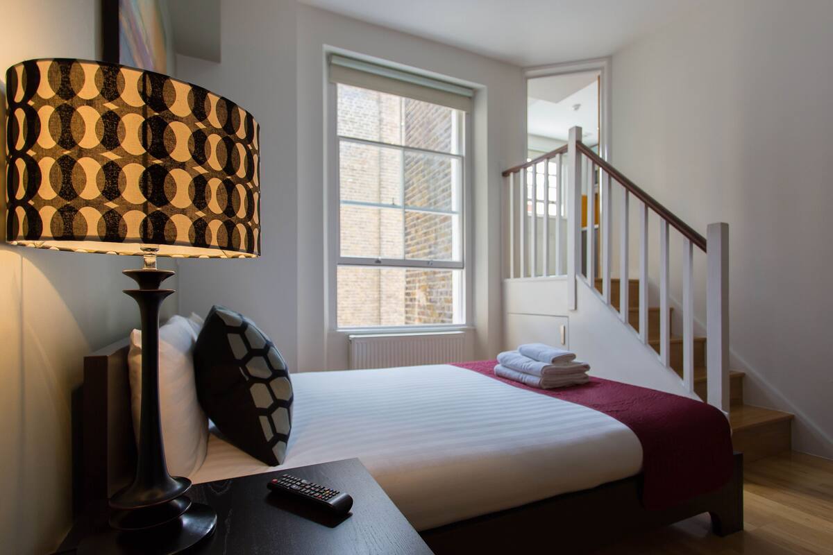 A hotel room with a bed, a nightstand with a lamp, a window, and a staircase.
