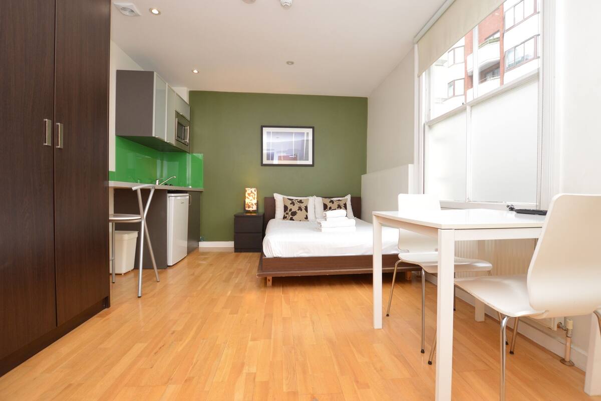 A compact room with a bed, a desk, a chair, a small kitchenette, and a window.