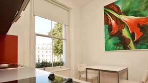 A modern kitchen with a black induction cooktop, white cabinets, and a window with a view of buildings and trees. There is a white table and two chairs in the room. - Princes Square Serviced Apartments by Concept Apartments (London)