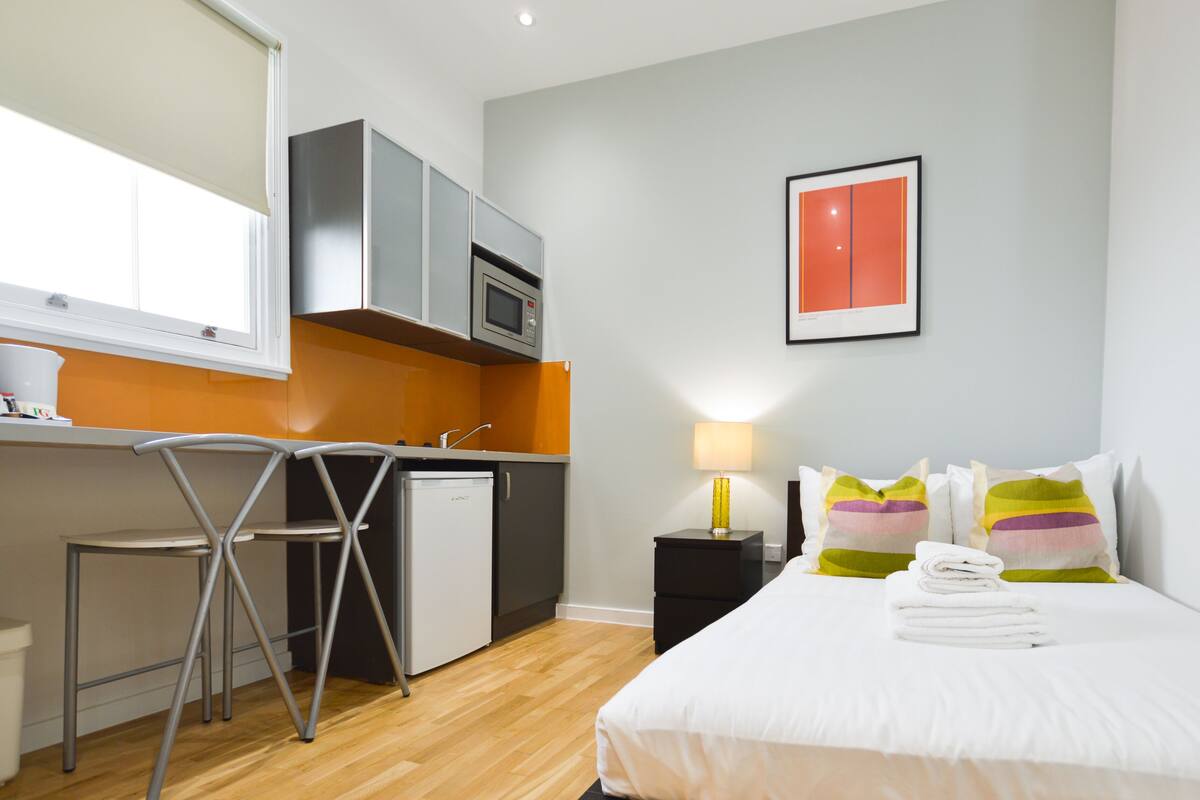 A compact hotel room with a kitchenette, a bed with colorful pillows, a bedside lamp, and a framed picture on the wall.