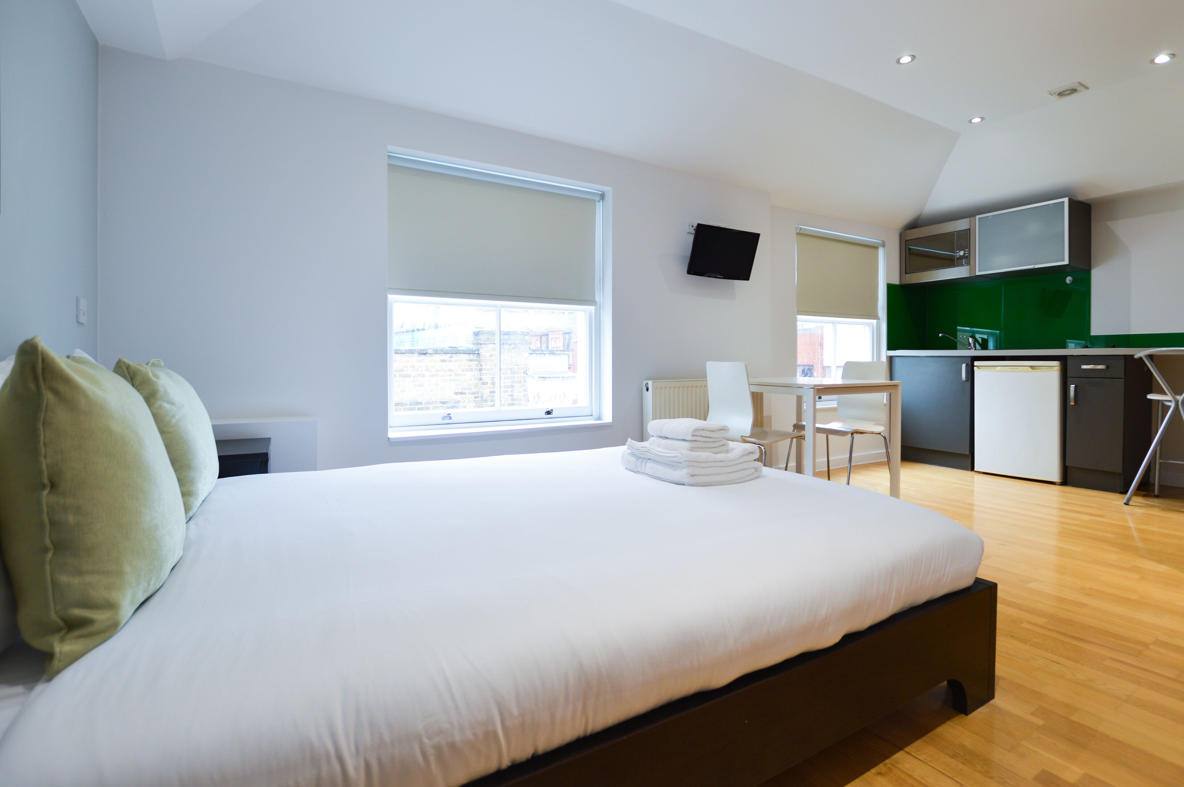 A modern hotel room with a large bed, a desk, a chair, and a kitchenette.