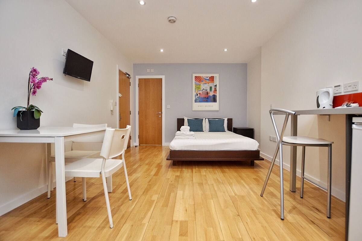 A modern hotel room with a wooden floor, a bed with white bedding, a desk with a chair, a TV mounted on the wall, and a poster on the wall.