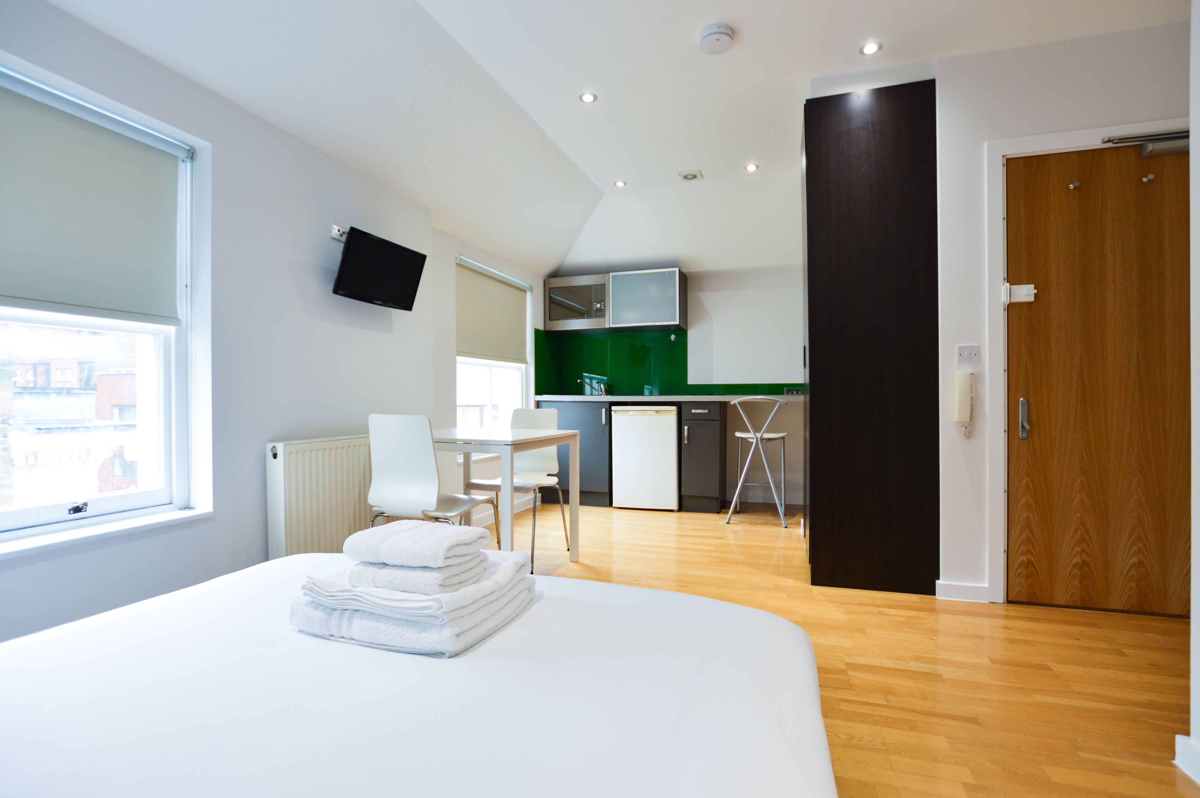 A modern hotel room with a bed, a desk, a chair, a small kitchenette, and a TV mounted on the wall.