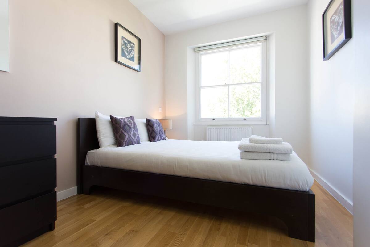 A bedroom with a bed, a dresser, a window, and framed artwork on the wall.