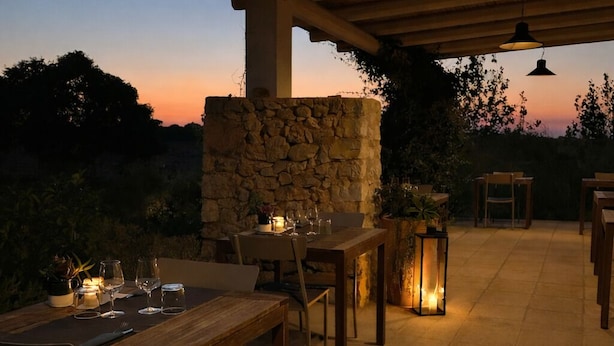 An outdoor patio with stone walls, wooden tables, and chairs, set against a sunset backdrop. - I Mulicchi (Specchia)