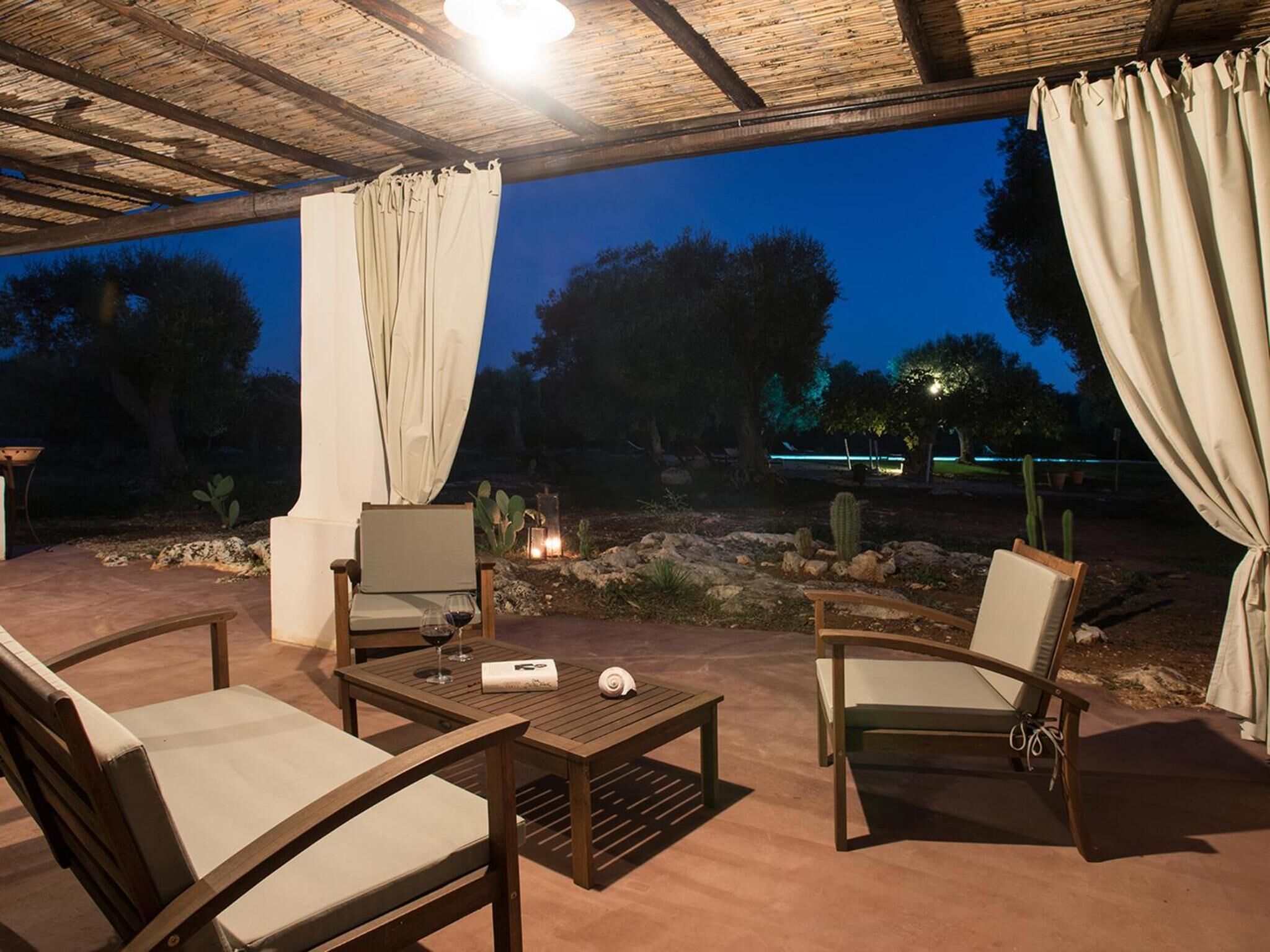 An outdoor patio area with two wooden chairs and a table, surrounded by a wooden pergola and draped curtains.