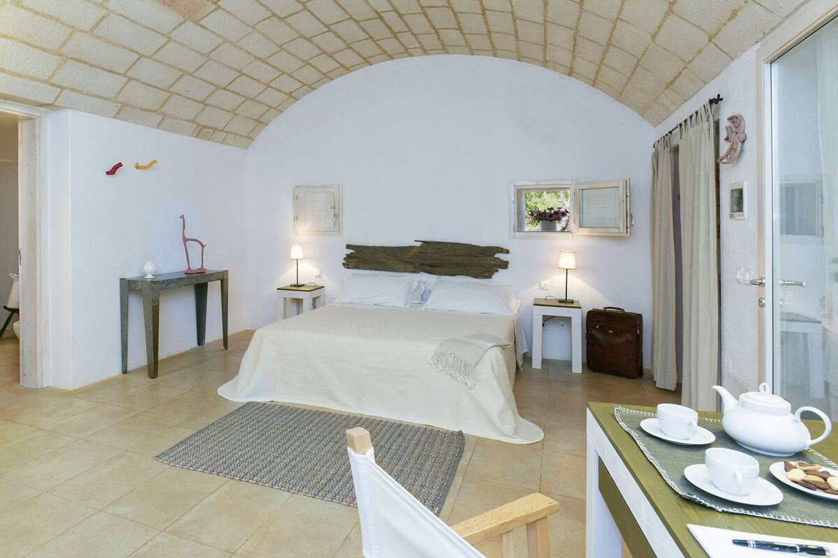 Un dormitorio con cama, mesitas de noche, un escritorio con juego de té y una ventana con cortinas.