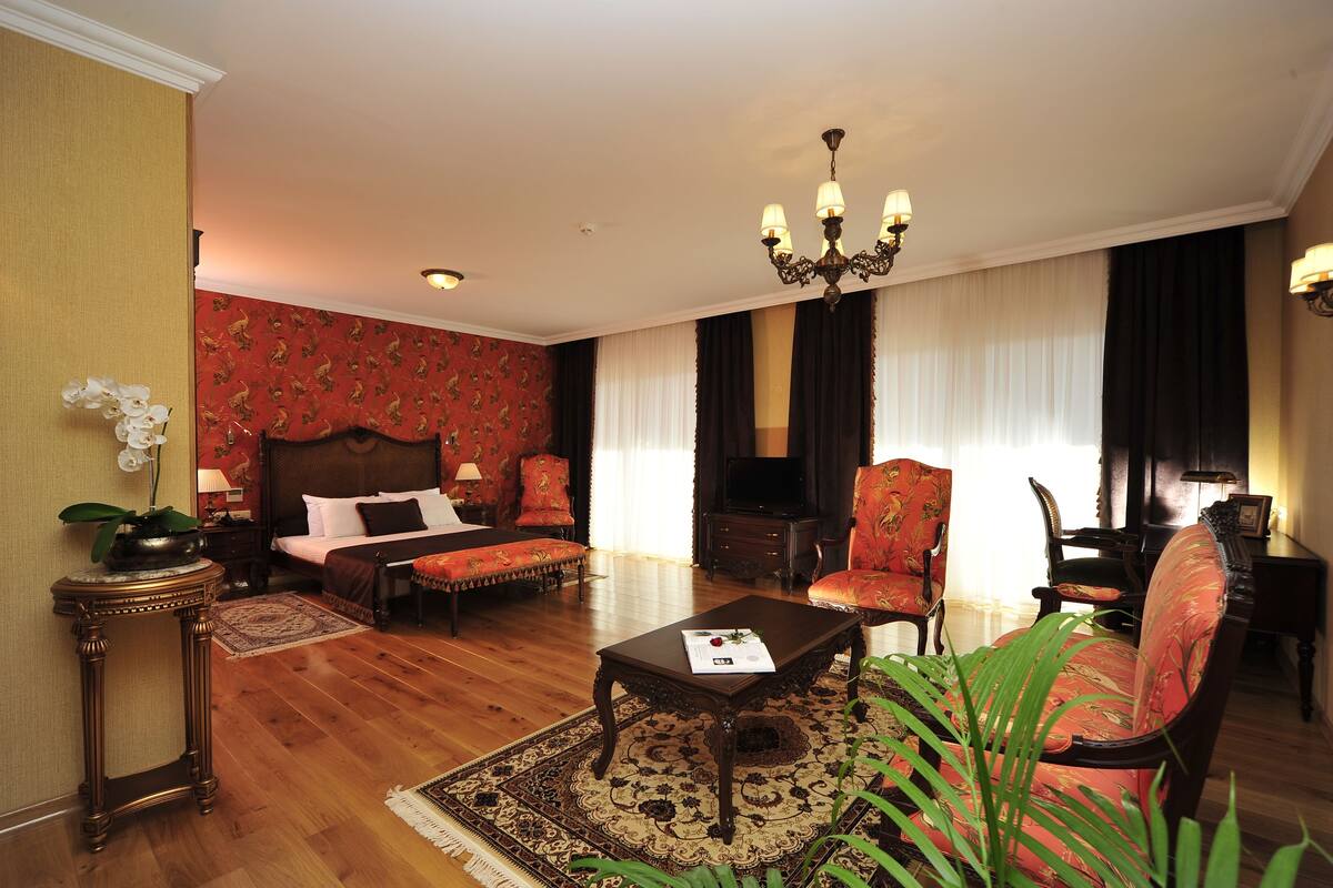 Una habitación de hotel con una cama grande, una zona de estar con sofá y sillones, una mesa de centro, una araña y un televisor.