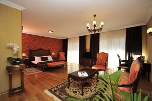 Una habitación de hotel con una cama grande, una zona de estar con sofá y sillones, una mesa de centro, una araña y un televisor.