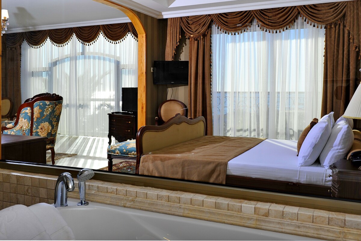 Habitación de hotel con un espejo grande, una cama con ropa blanca, un cabecero de madera, una mesita de noche con una lámpara y vistas de la habitación a través de cortinas de gasa.