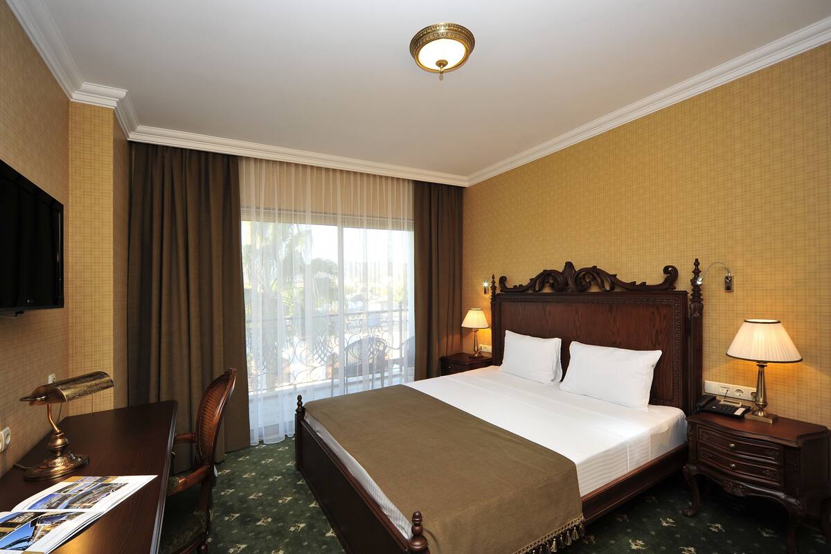 Habitación de hotel con una cama grande, un escritorio con lámpara, una silla, un televisor y una ventana con cortinas.