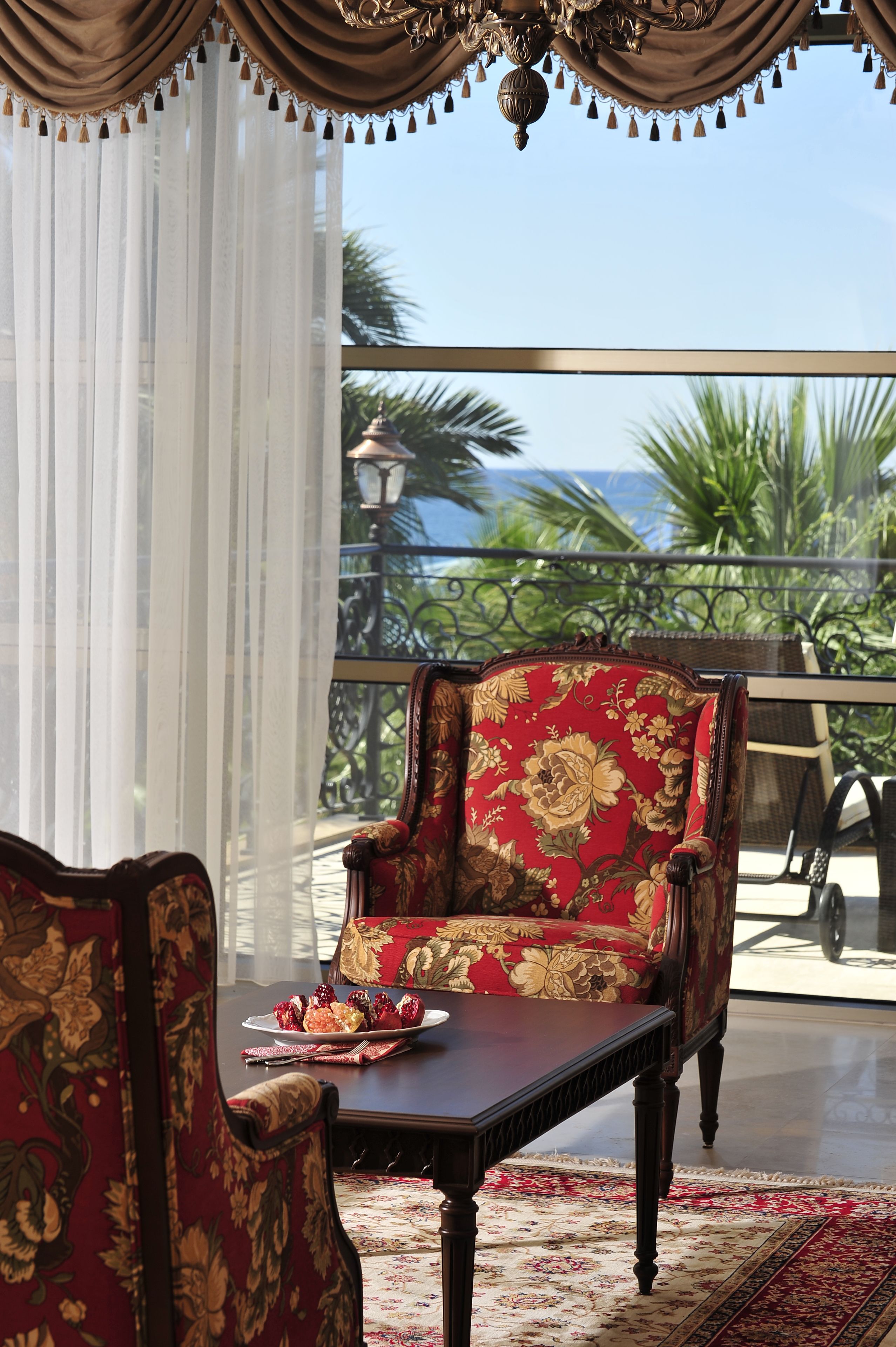 A balcony with a red floral chair and a table, a view of the ocean, and a palm tree in the background.