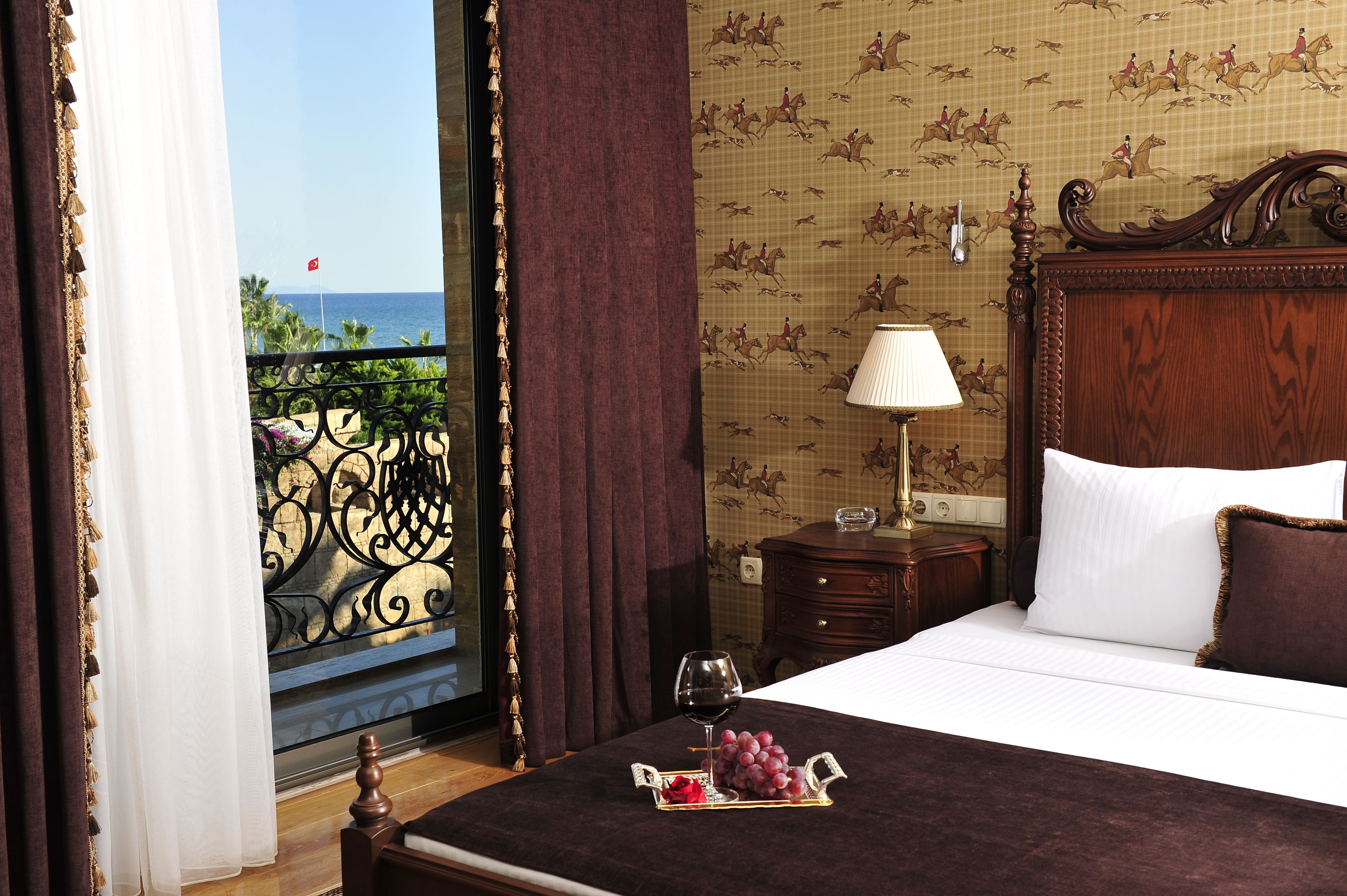 A hotel room with a bed, a bedside table with a lamp, a tray with grapes and a glass of wine, and a view of the sea through a balcony with a railing.