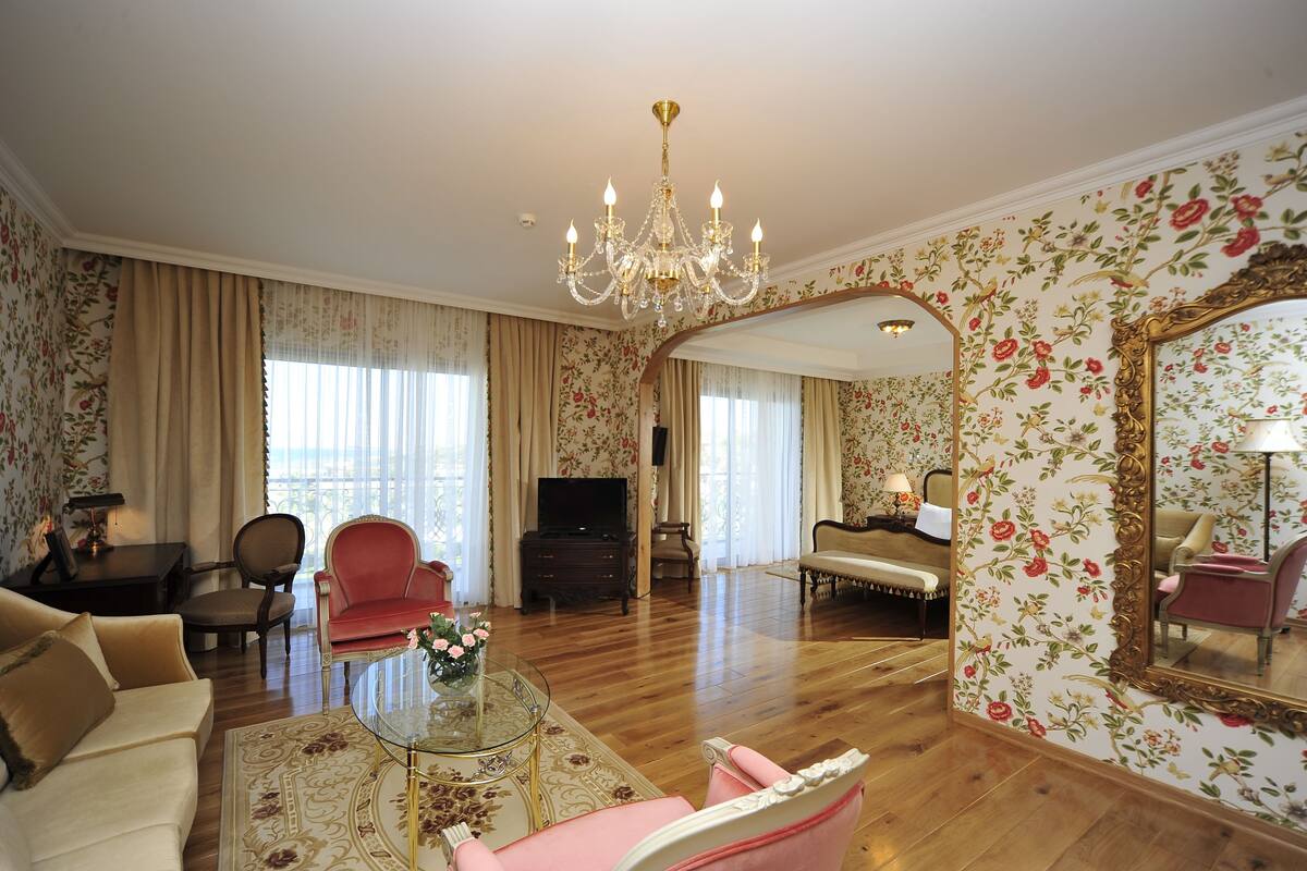 Un salón amplio con una araña, papel tapiz floral, un televisor y un espejo.