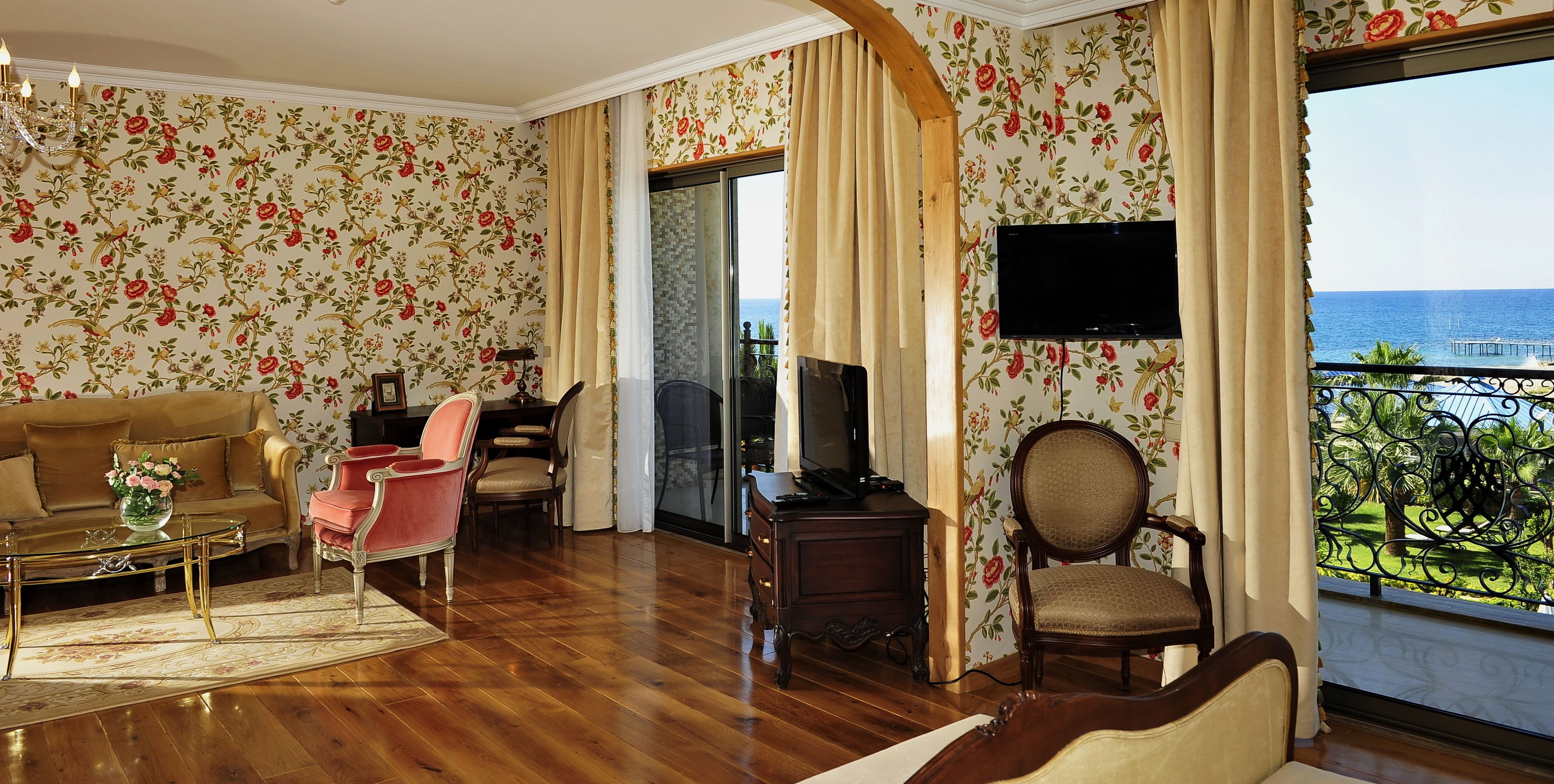 A living room with a sofa, chairs, a coffee table, and a TV. There is a balcony with a view of the sea.