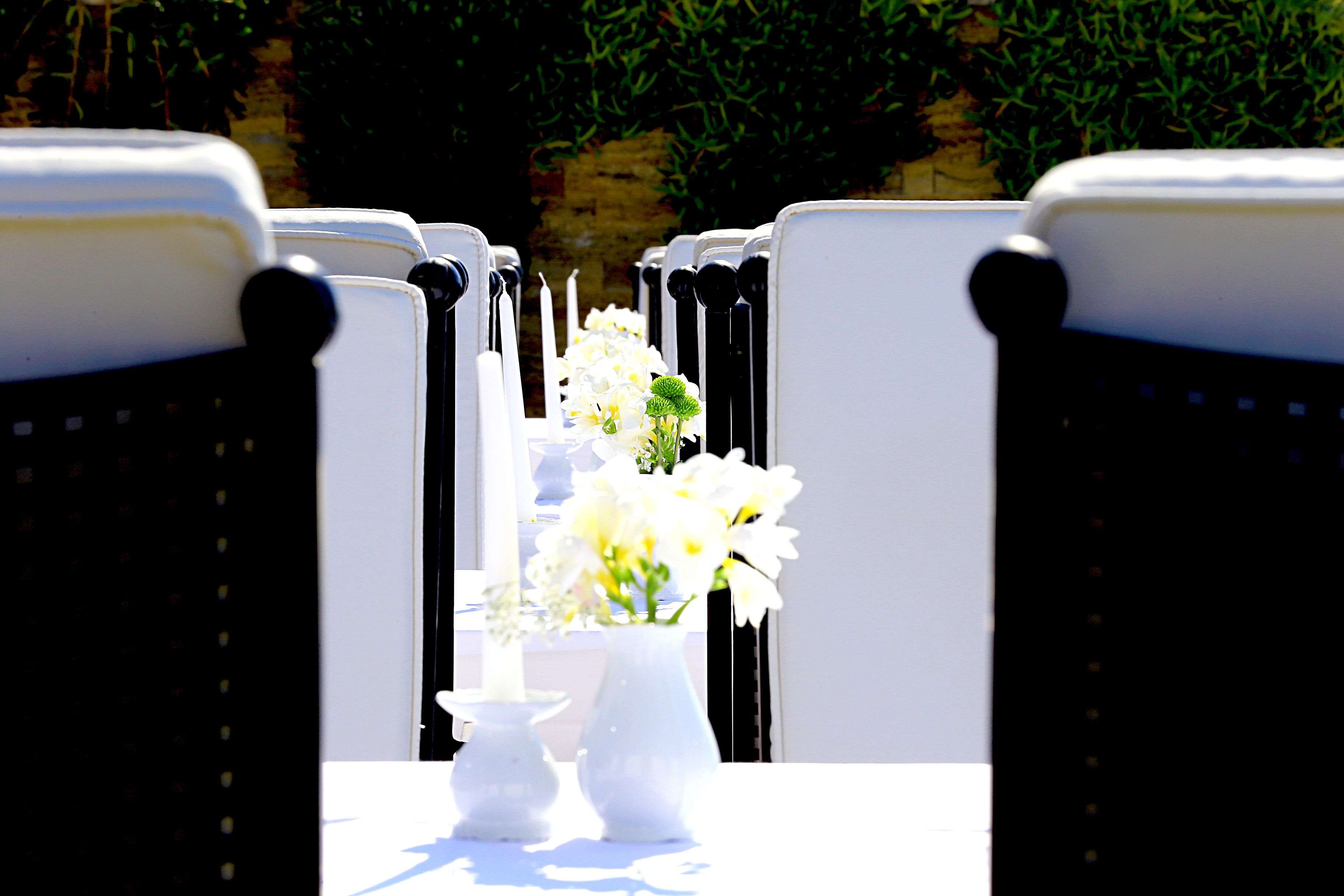 Une rangée de chaises blanches avec des accoudoirs noirs et une table blanche ornée d’un vase rempli de fleurs blanches.