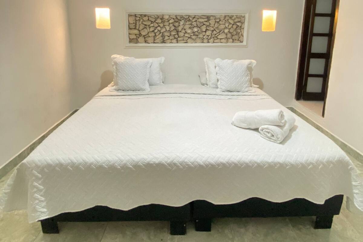 Una cama bien hecha con sábanas y almohadas blancas, una lámpara fijada a la pared y un adorno de pared de piedra.