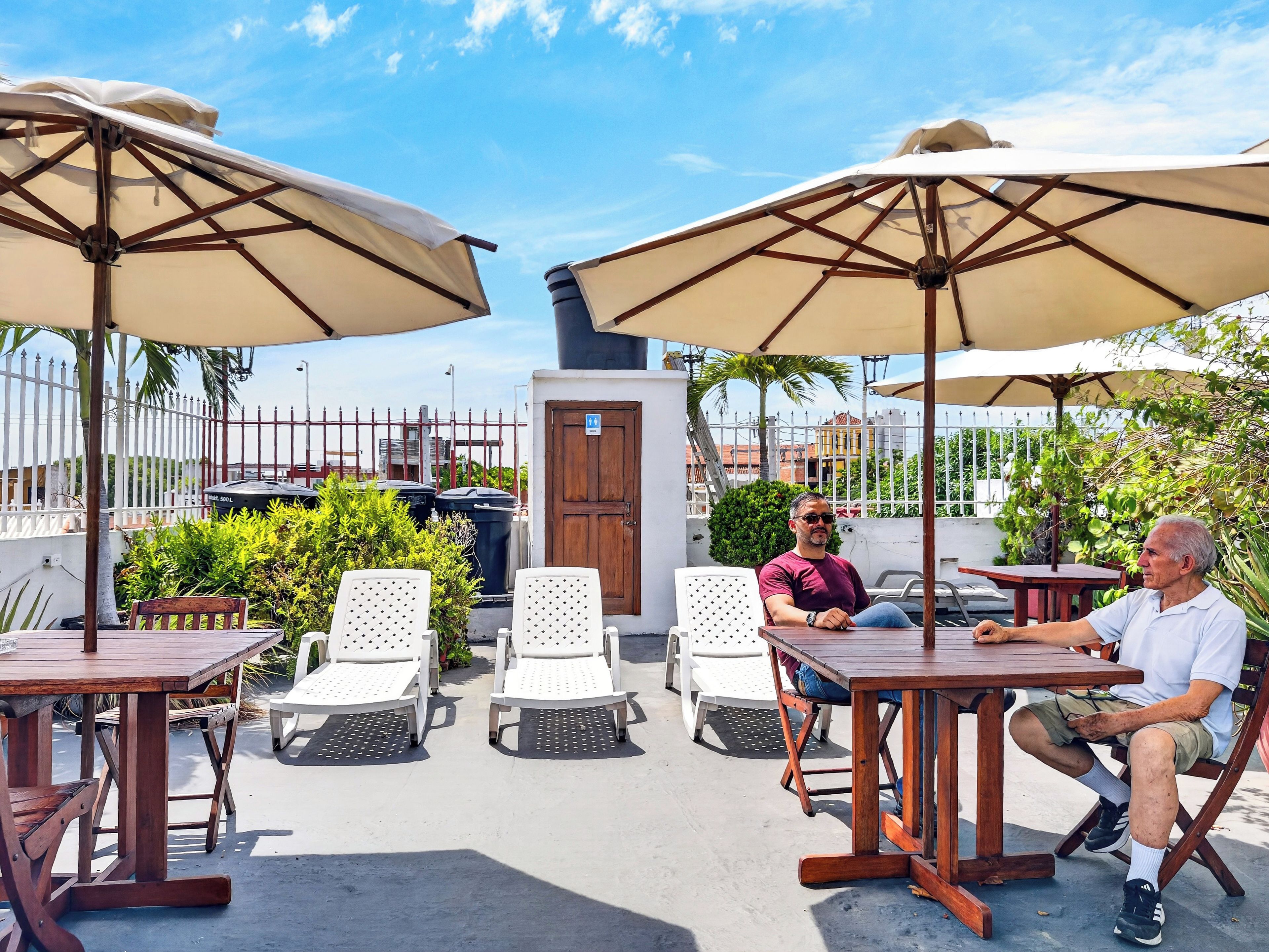 An outdoor seating area with wooden tables and chairs, shaded by large umbrellas, and a view of the cityscape in the background.