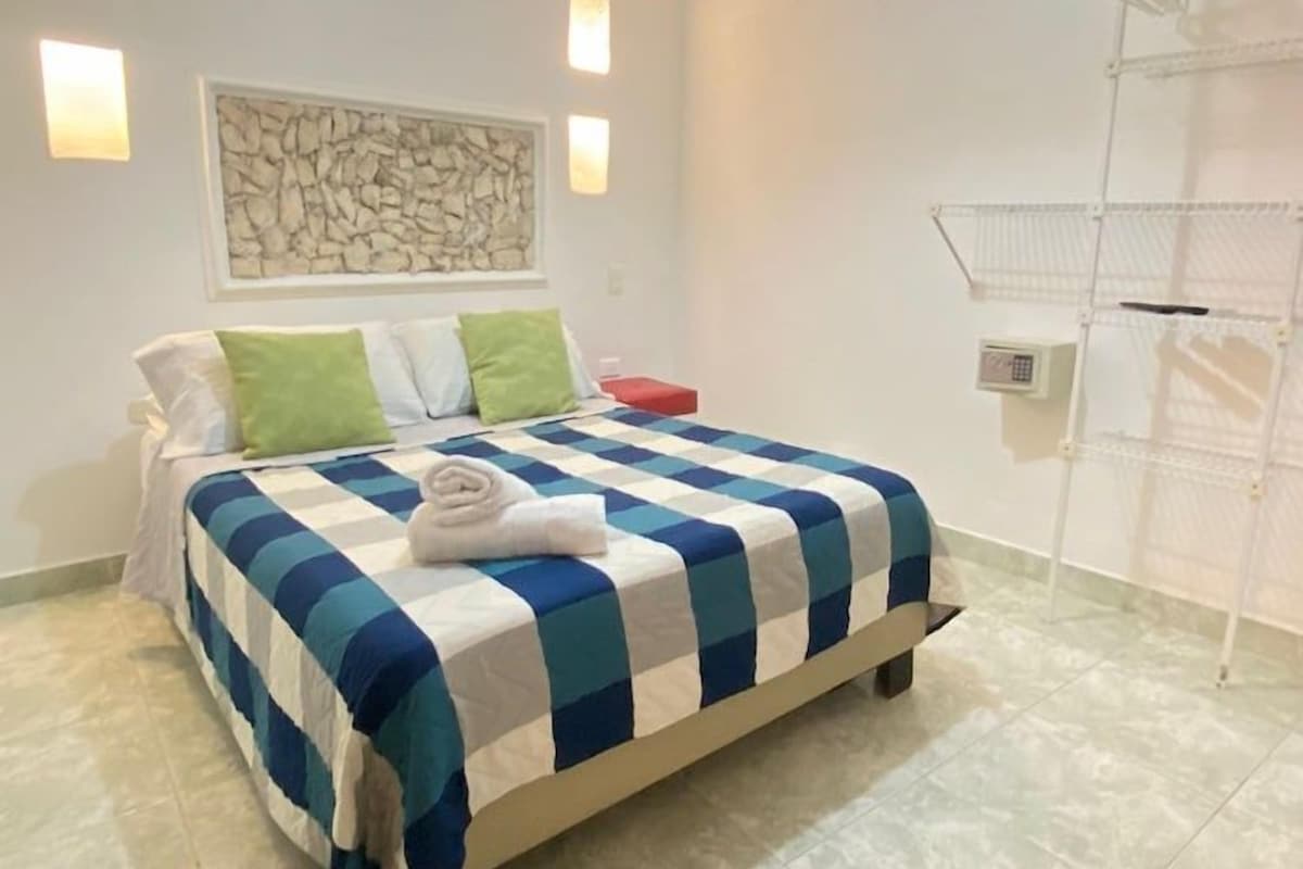A hotel room with a bed, a wall-mounted air conditioner, a ceiling light, and a wall-mounted shelf.