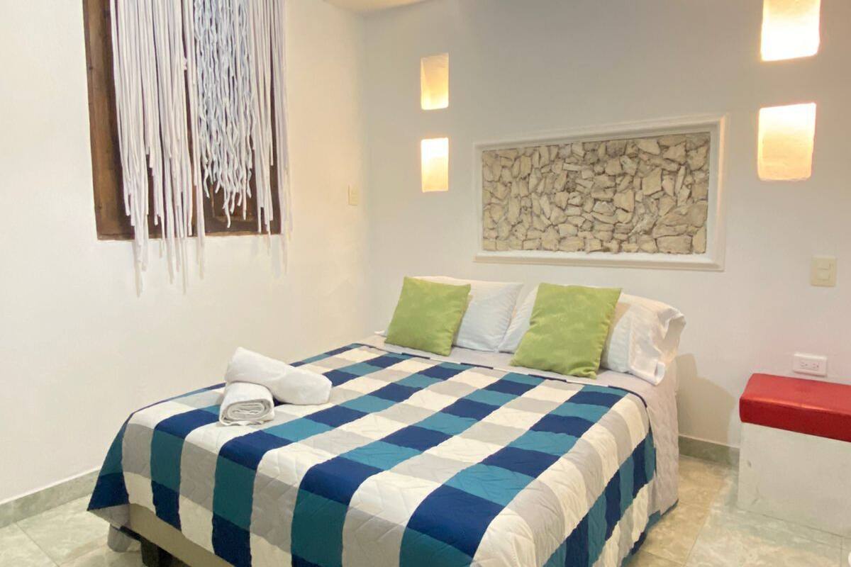 Una cama bien hecha con colcha azul y blanca a cuadros, almohada blanca y almohadones verdes decorativos. Arriba, una lámpara de pared y, abajo, una decoración de pared de piedra.